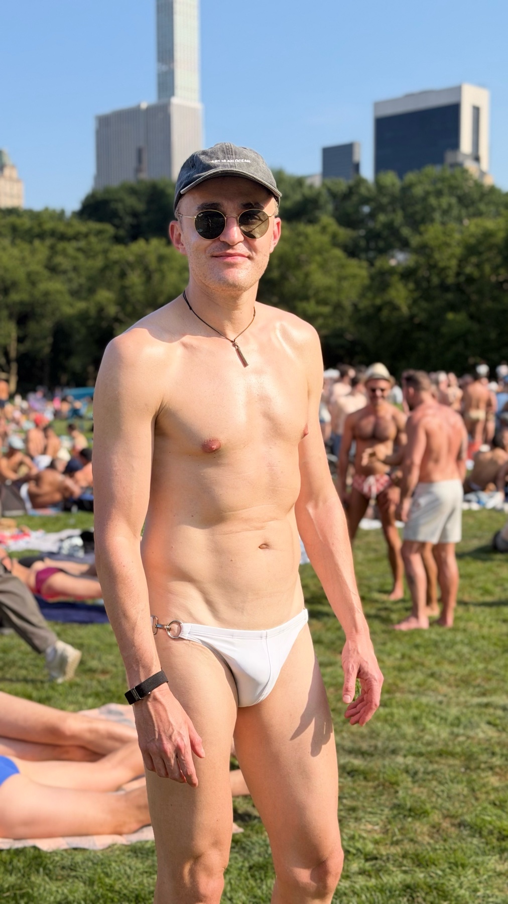 A shirtless man wearing sunglasses and a baseball cap poses for a photo in Central Park, with other people sunbathing in the background.