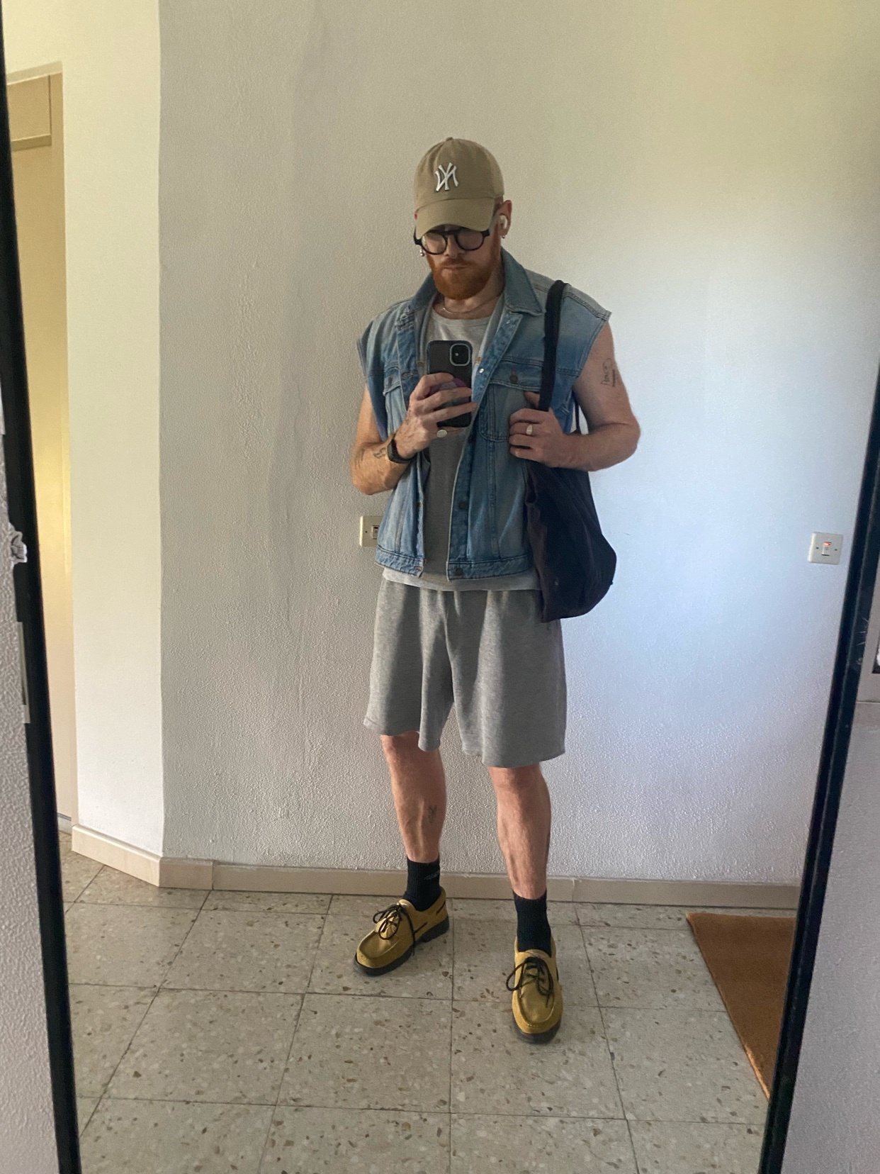 A man with red hair, a beard, and glasses poses in front of a mirror. He is wearing a denim vest over a t-shirt, grey shorts, and yellow sneakers.