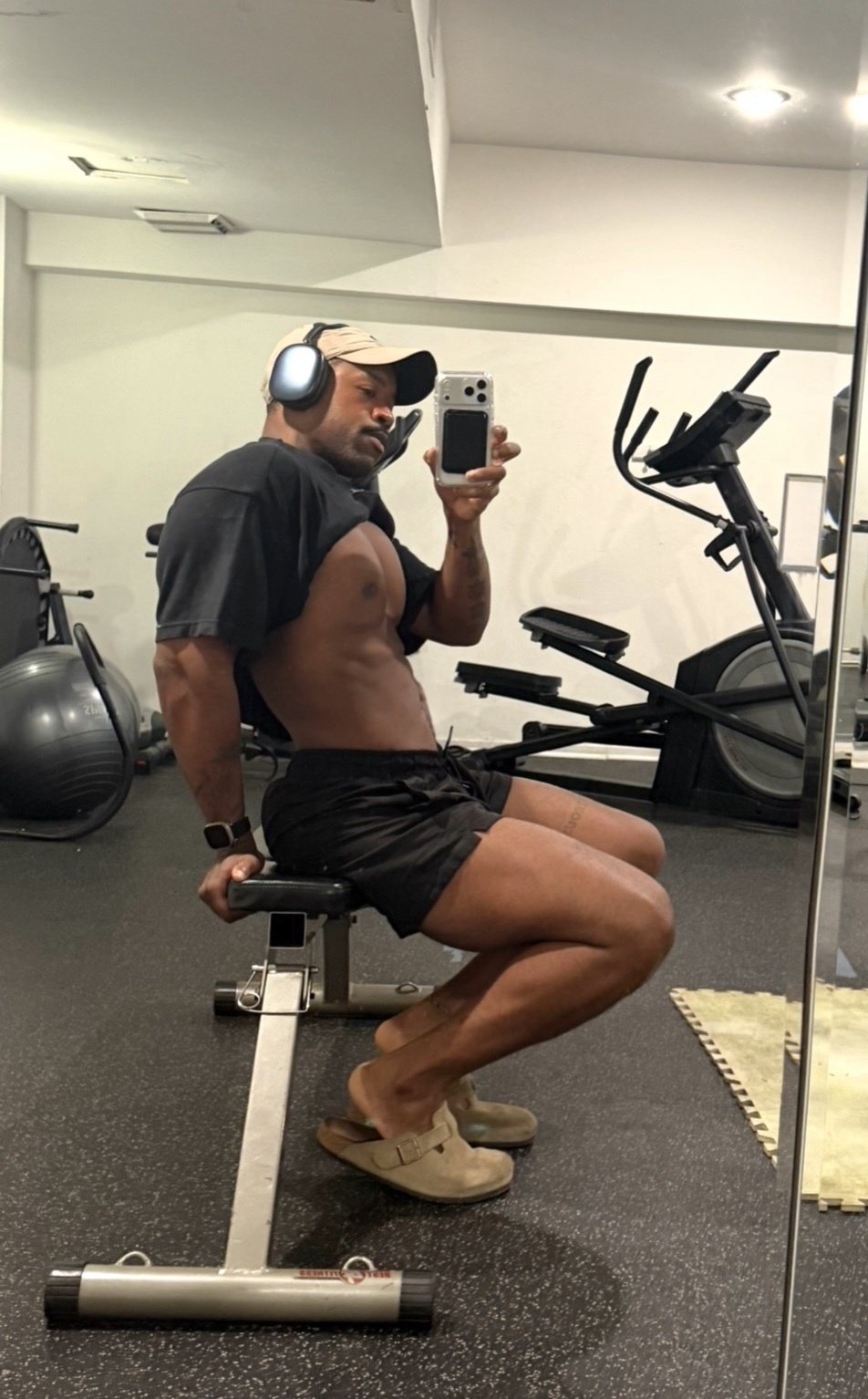 A man with a muscular physique takes a selfie in a gym, wearing headphones and a backward baseball cap. He is shirtless, showing his abs and biceps, and is wearing black shorts and clogs.