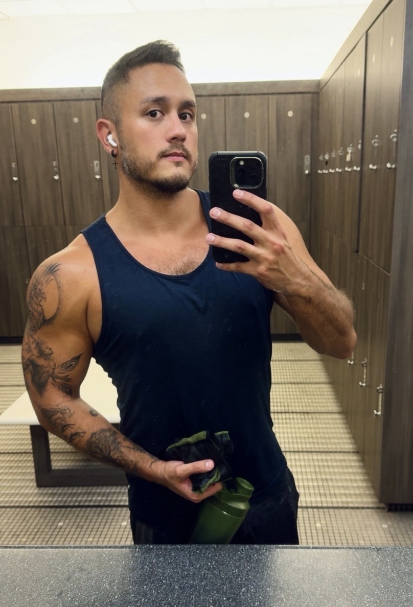A man with tattoos on his arm and stubble poses for a selfie in a gym locker room. He is wearing a dark tank top and holding a green water bottle.