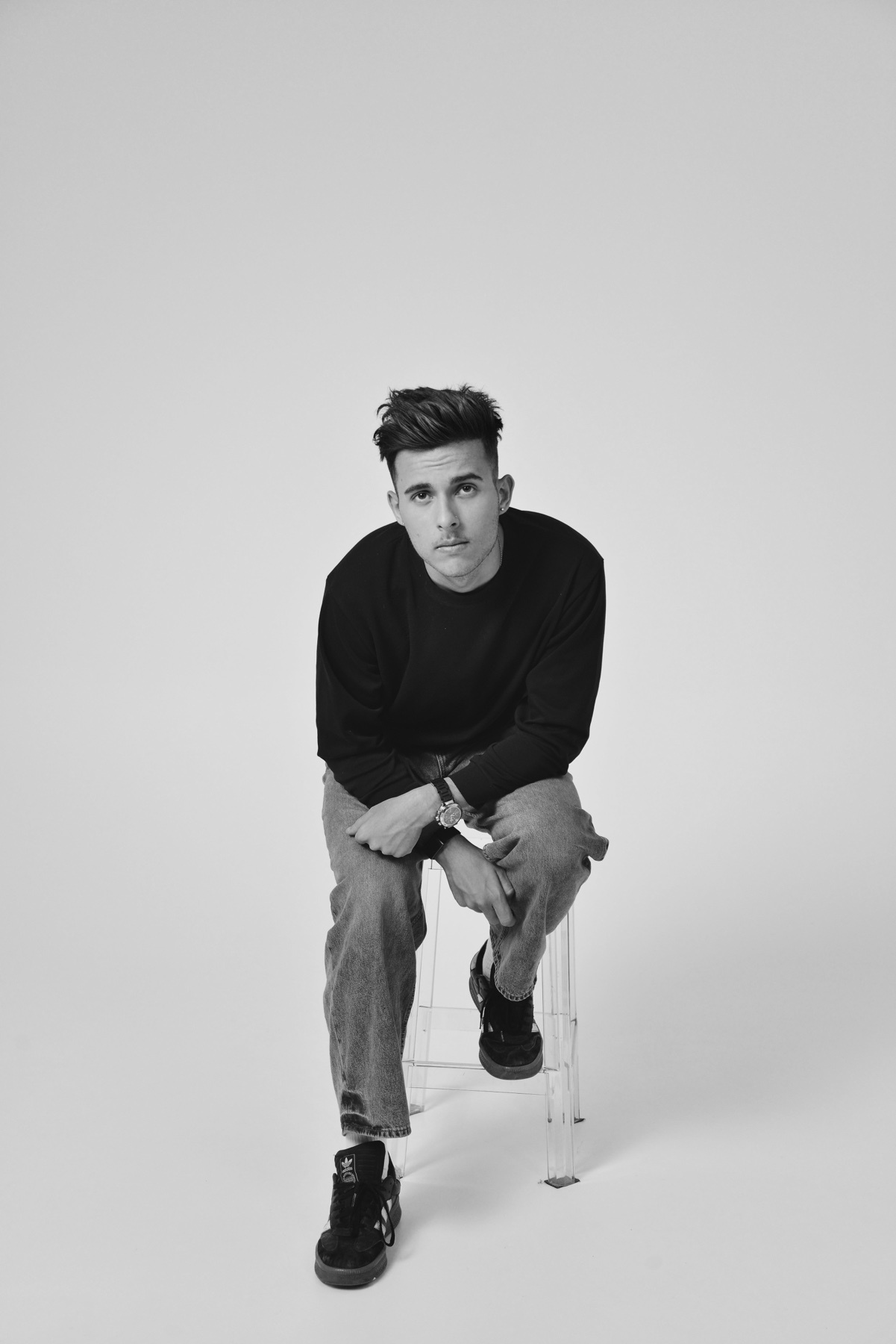 A black and white studio portrait of a man. He is sitting on a clear stool, looking directly at the camera.