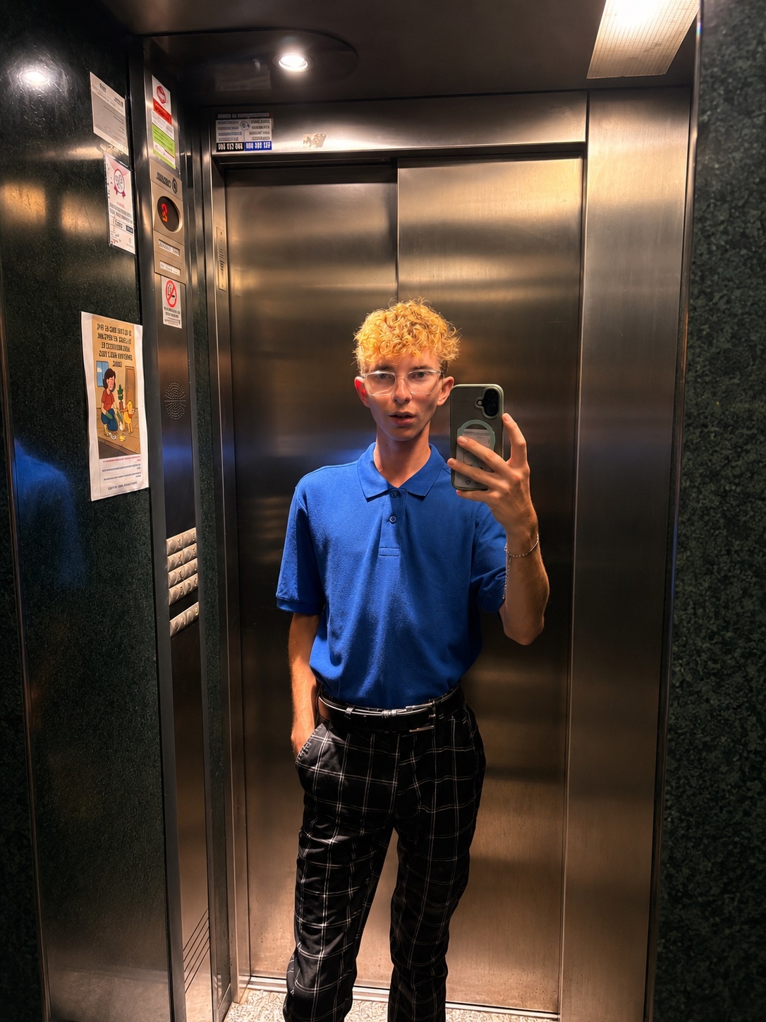 A young man with curly blonde hair and glasses takes a selfie in an elevator. He is wearing a blue polo shirt and plaid pants.