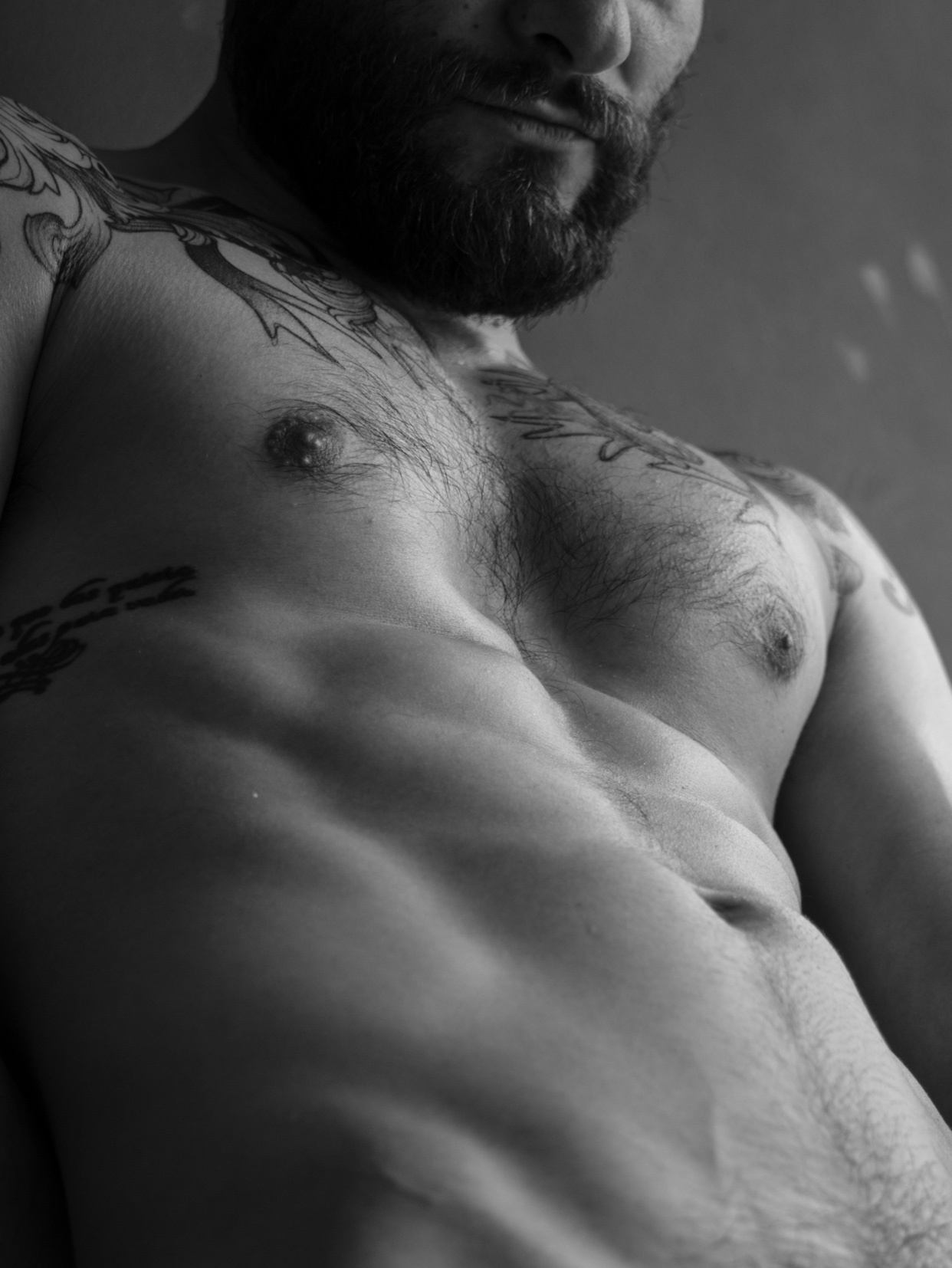 A black and white photo of a man with tattoos and a beard. The man is shirtless, showcasing his muscular physique.