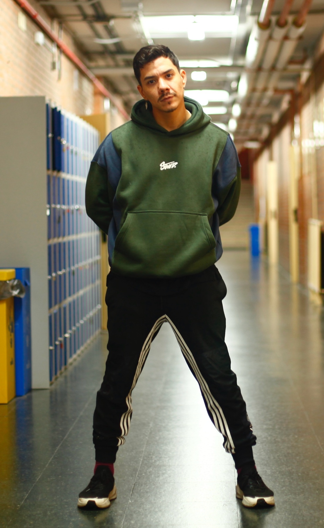 A man stands in a hallway, posing with his hands behind his back. He is wearing a green and blue color-blocked hoodie, black track pants with white stripes, and black sneakers.
