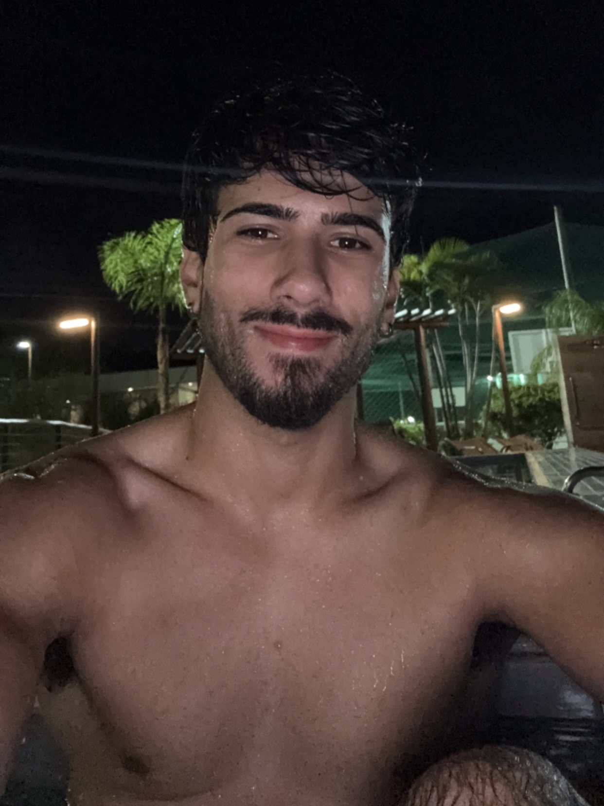 A shirtless man with dark hair, a beard, and stubble smiles at the camera at night. He appears to be in a pool or hot tub, with water droplets visible on his skin.