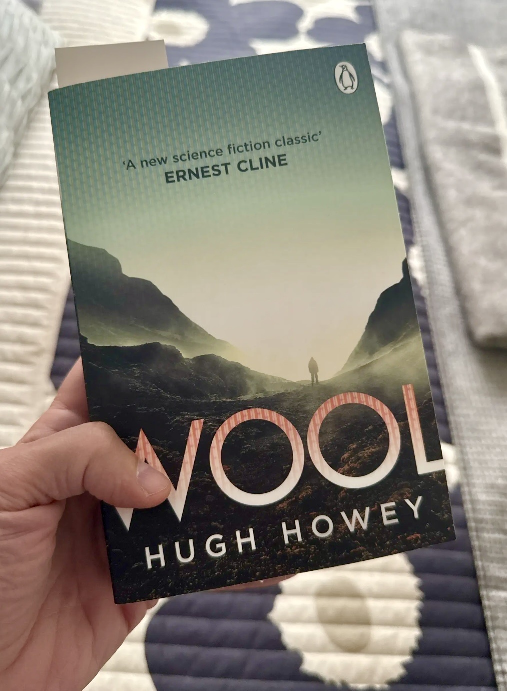 A hand holds the book "Wool" by Hugh Howey, featuring a dramatic landscape with a lone figure silhouetted against mountains.
