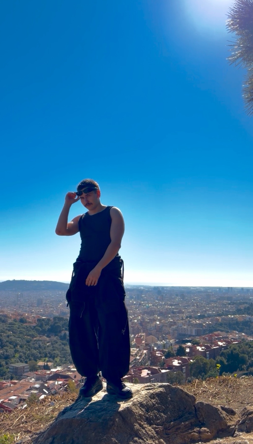 A man stands on a rock overlooking a city on a clear, sunny day. He is wearing a black tank top, black pants, a black baseball cap, and sunglasses, and he is shielding his eyes from the sun.