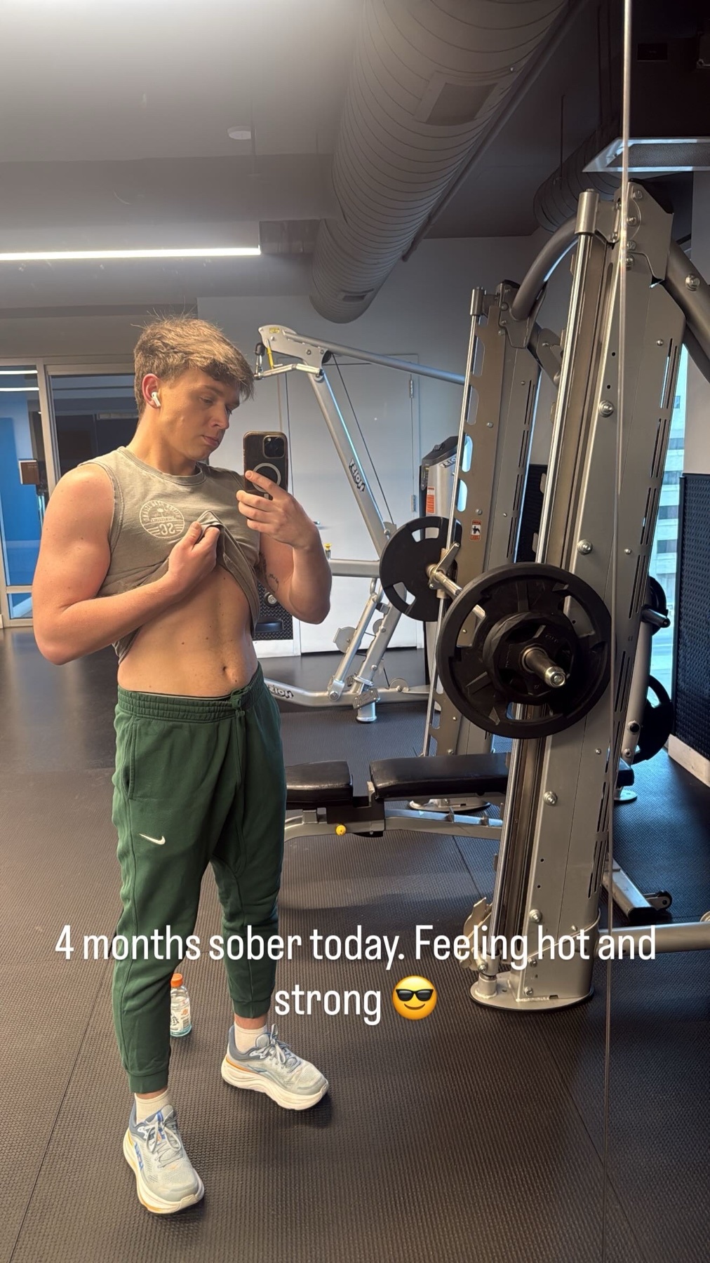 A man in athletic wear poses for a mirror selfie in a gym, showing off his physique and celebrating four months of sobriety.