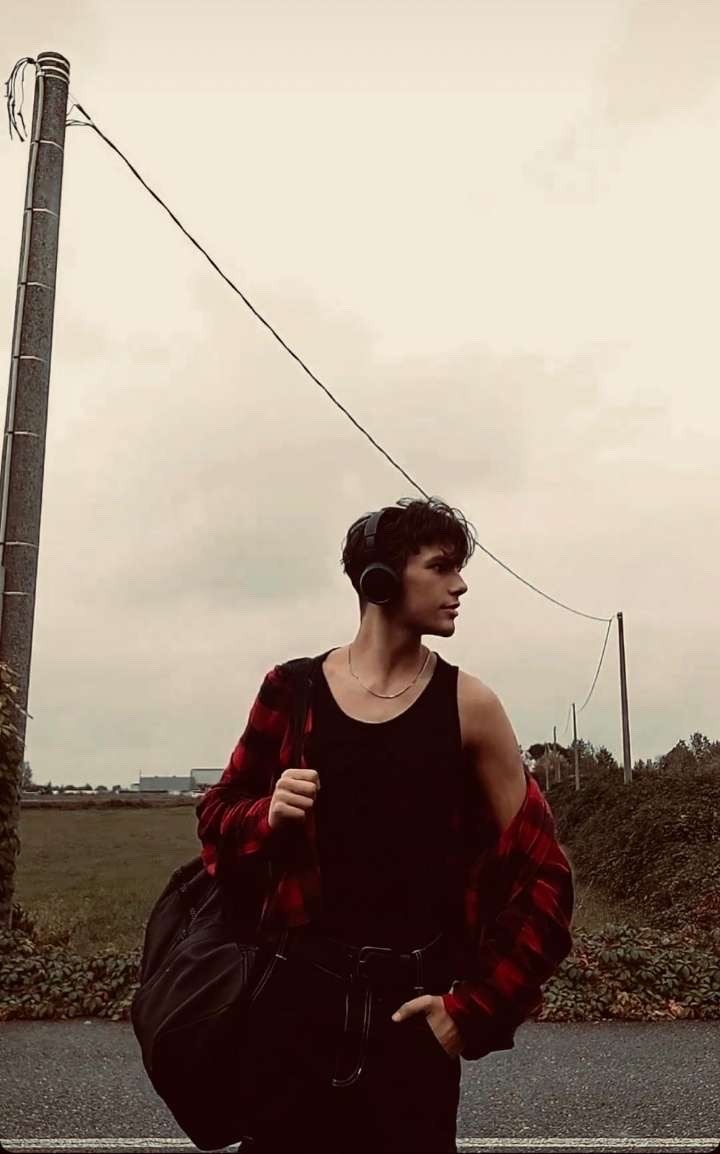 A young man wearing a black tank top and an open red and black flannel shirt stands outside with headphones on, holding a duffel bag.