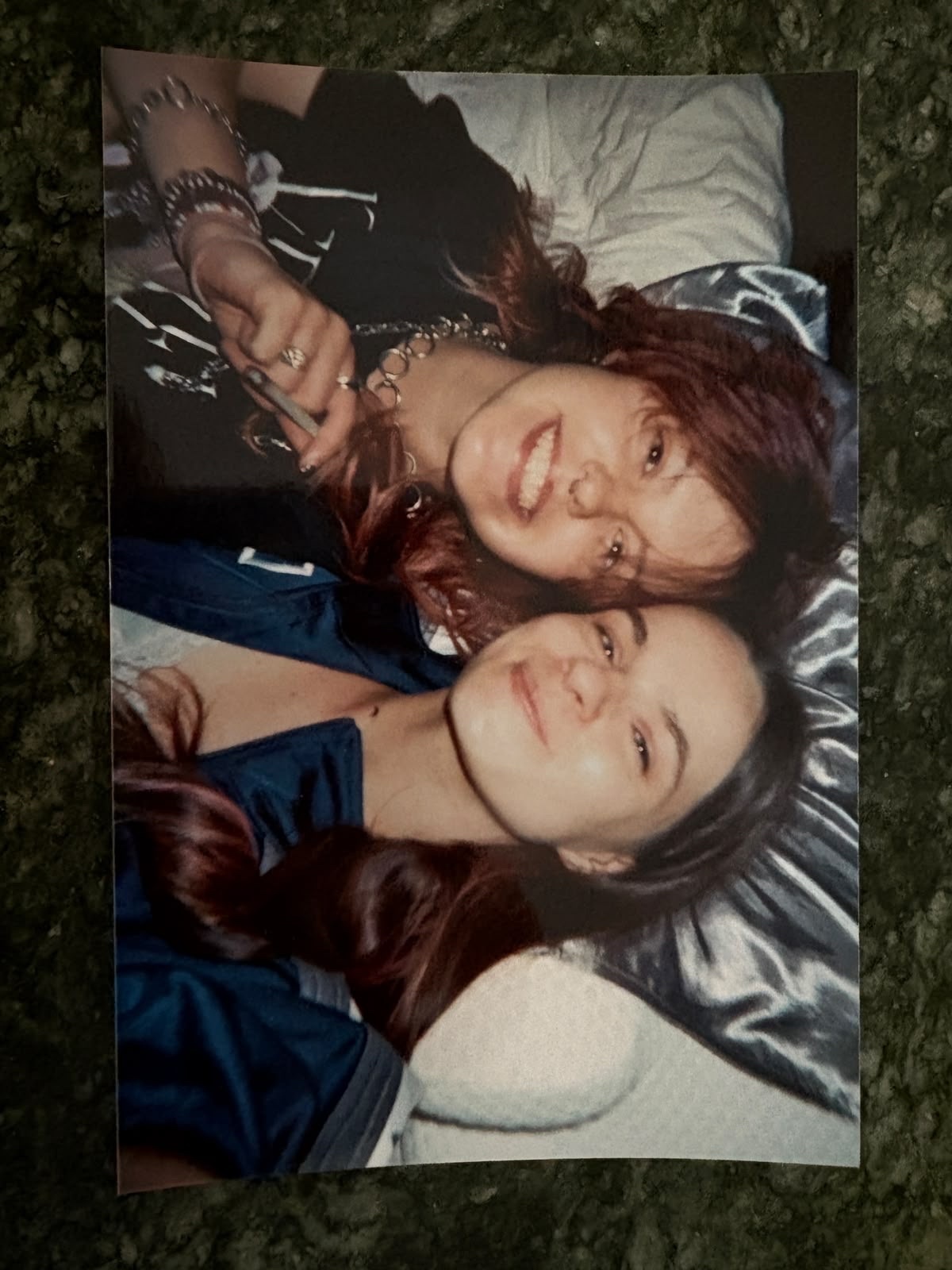 Two women are lying down and smiling at the camera. The woman in the foreground has long, dark hair with some reddish highlights and is wearing a blue jacket. The woman behind her has long, wavy, dark red hair and is wearing a black t-shirt with jewe