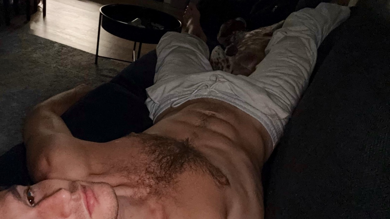A man is lying shirtless on a couch, with white pants. A dog is also present in the background.