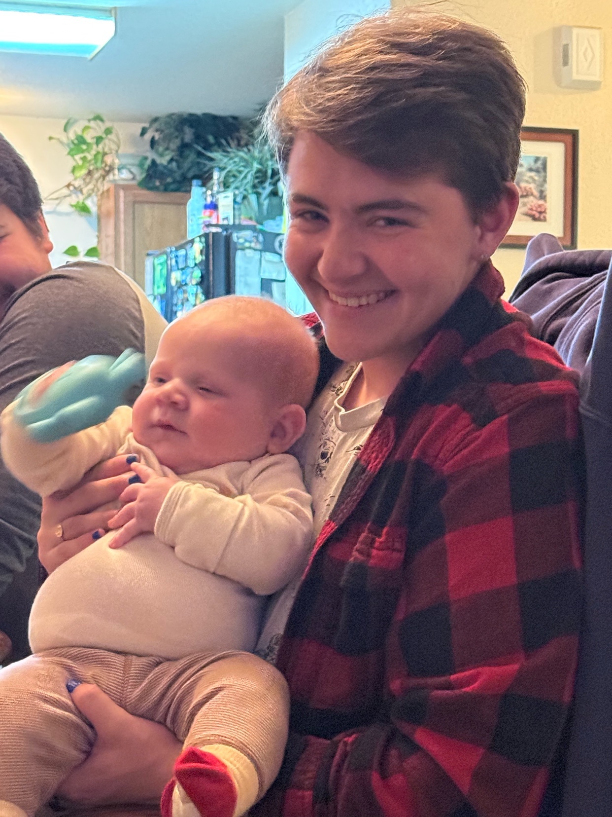 A person with short brown hair smiles while holding a baby. The person is wearing a red and black plaid shirt, and the baby is wearing a cream-colored onesie and tan pants.