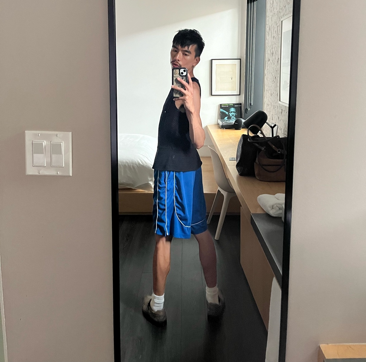A man takes a mirror selfie, showcasing his outfit. He is shirtless, wearing a tank top and blue shorts.