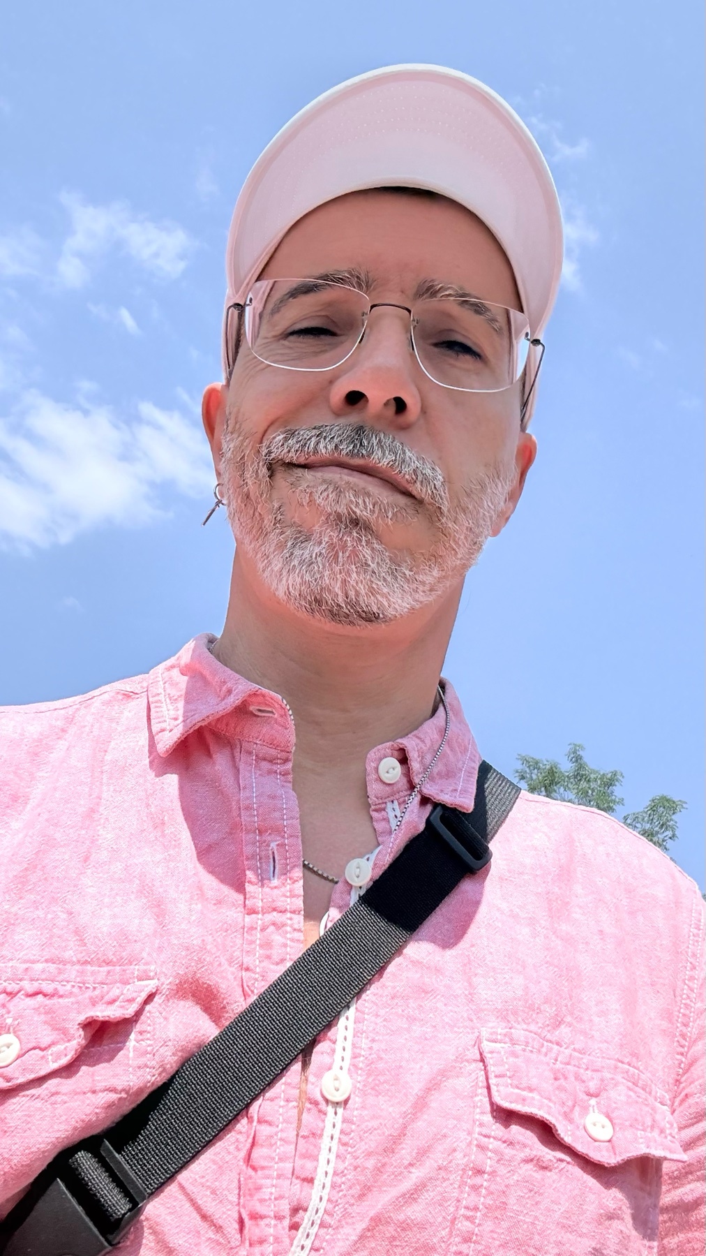 A man with glasses, a beard, and stubble takes a selfie outdoors under a blue sky. He is wearing a pink shirt and a white hat.