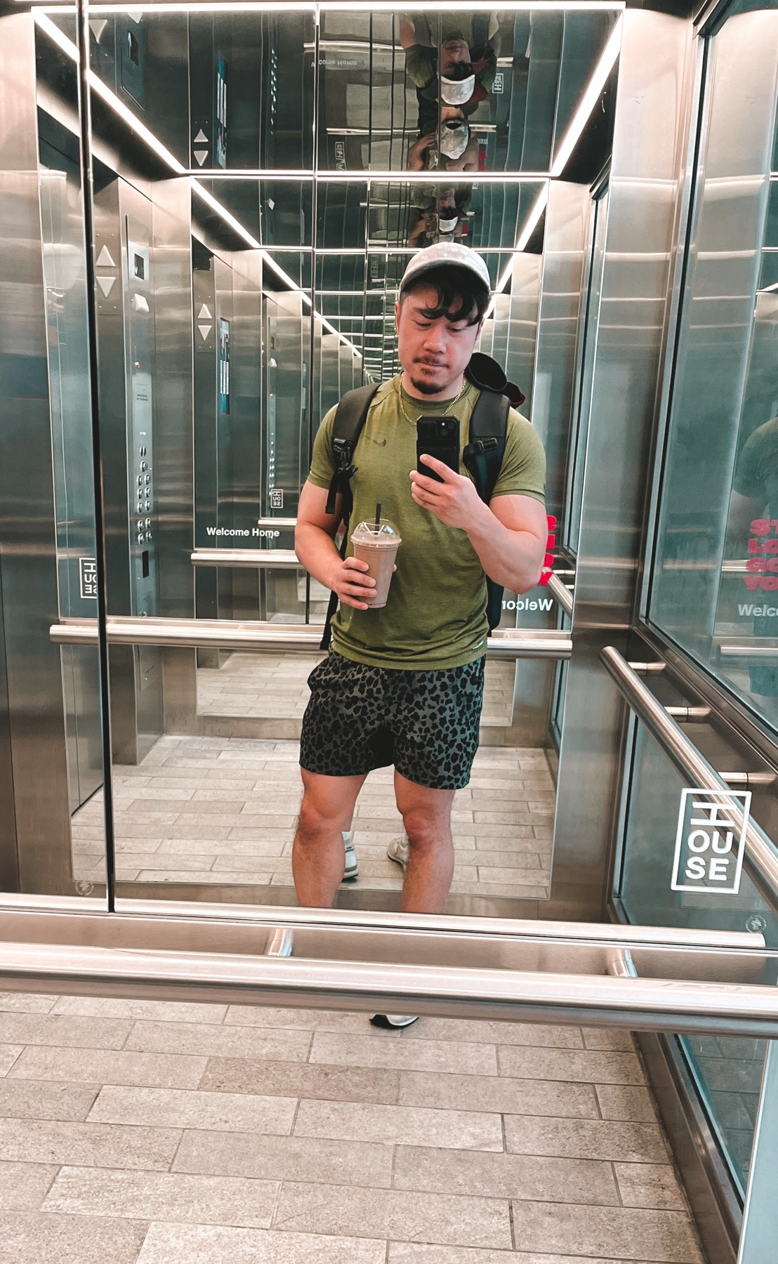 A man takes a mirror selfie in an elevator, holding a drink and wearing leopard print shorts.