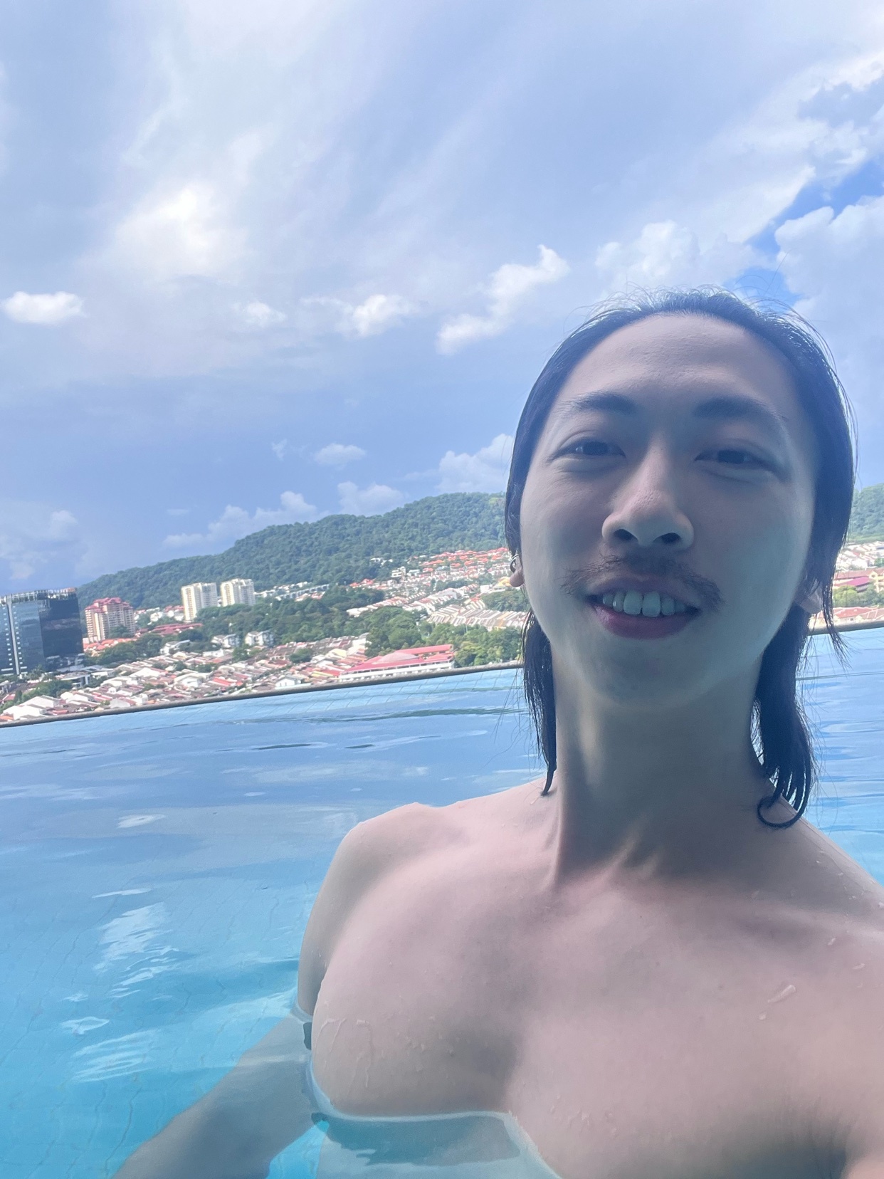 A man with a mustache and long dark hair takes a selfie in an infinity pool overlooking a cityscape.
