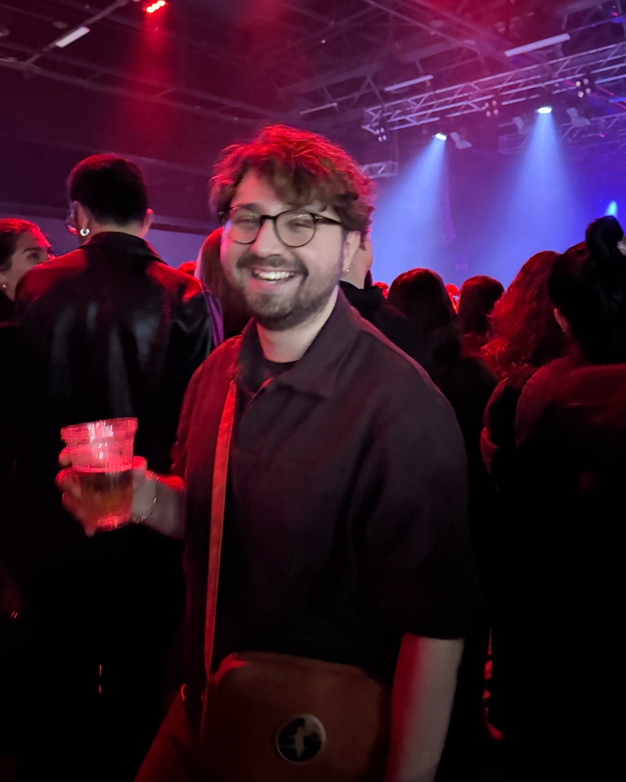 A man smiles while holding a drink at a party. He is surrounded by other people in a dimly lit indoor setting.