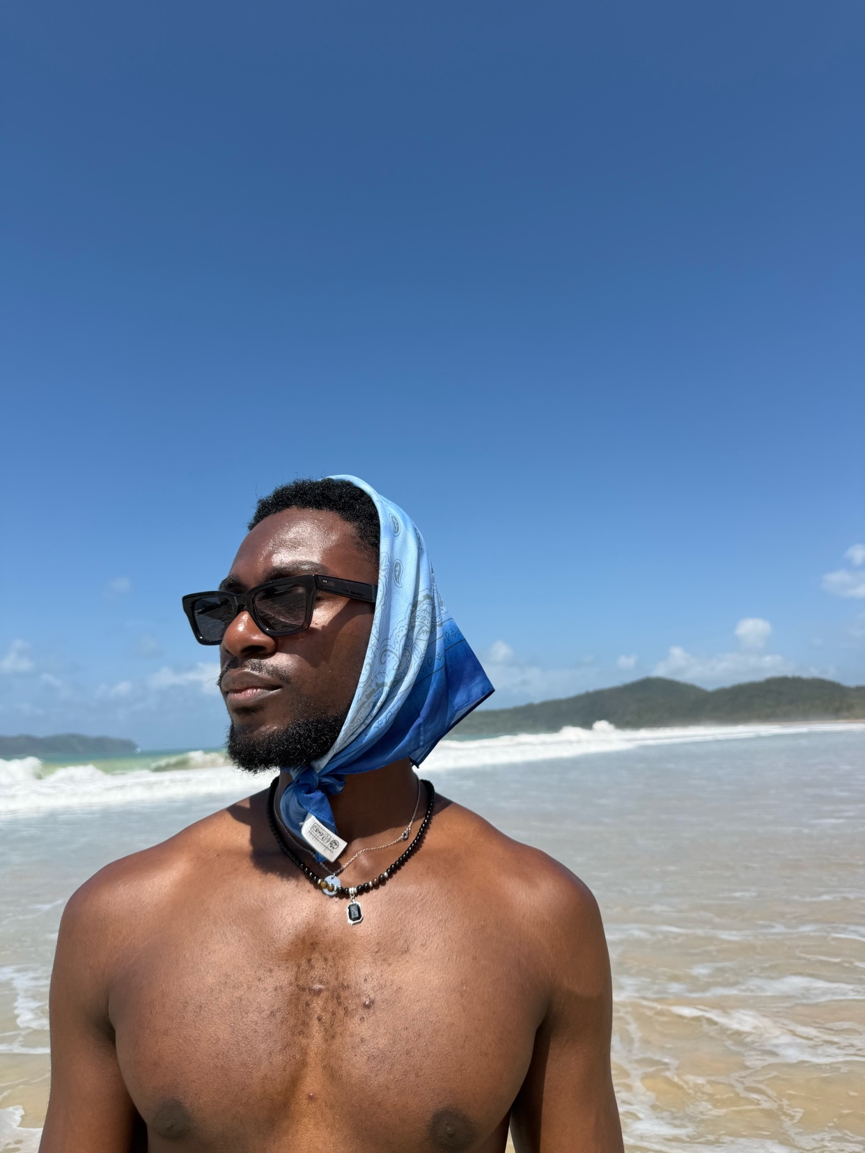 A man stands shirtless on a beach, wearing sunglasses and a headscarf. The photo captures a sunny day with the ocean and sky in the background.