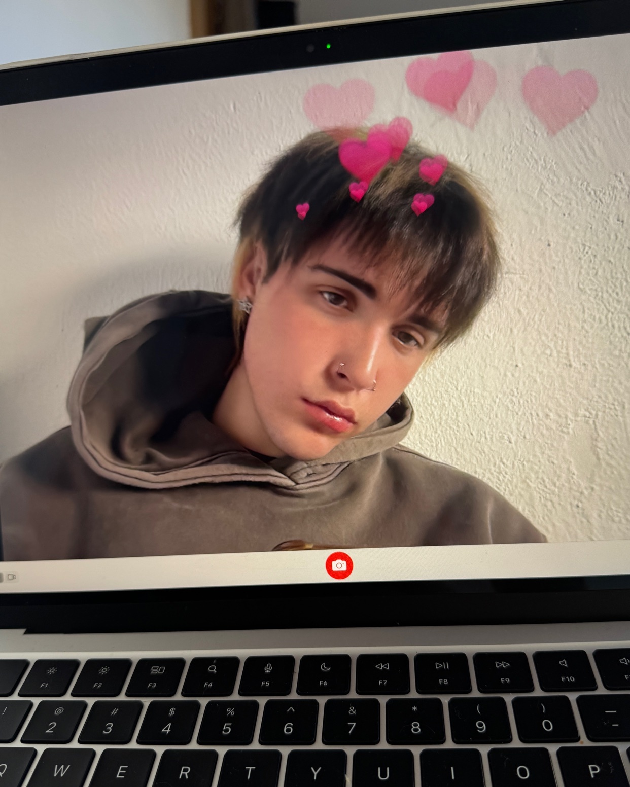 A young man poses for a selfie on a laptop screen. He is wearing a hoodie and has a heart filter applied.