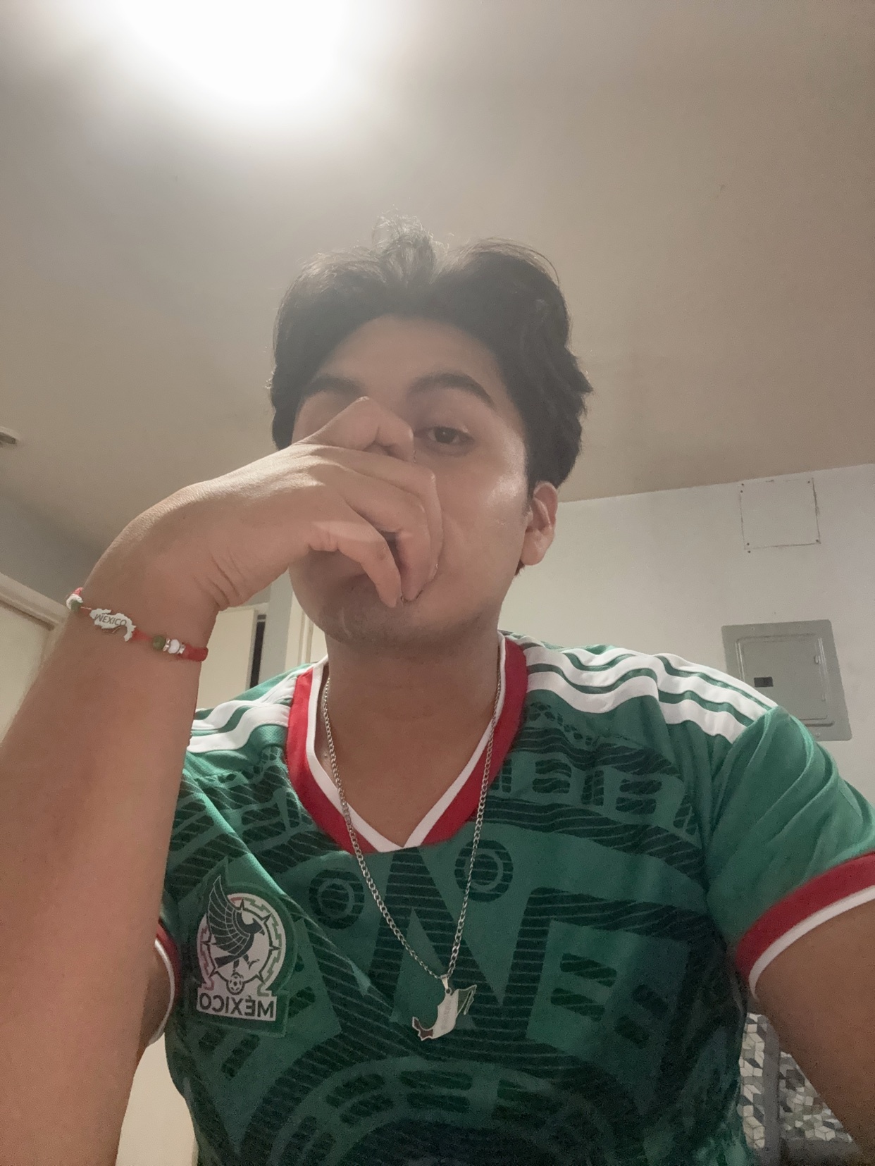 A young man wearing a Mexico national football team jersey poses for a selfie, covering his mouth with his hand.