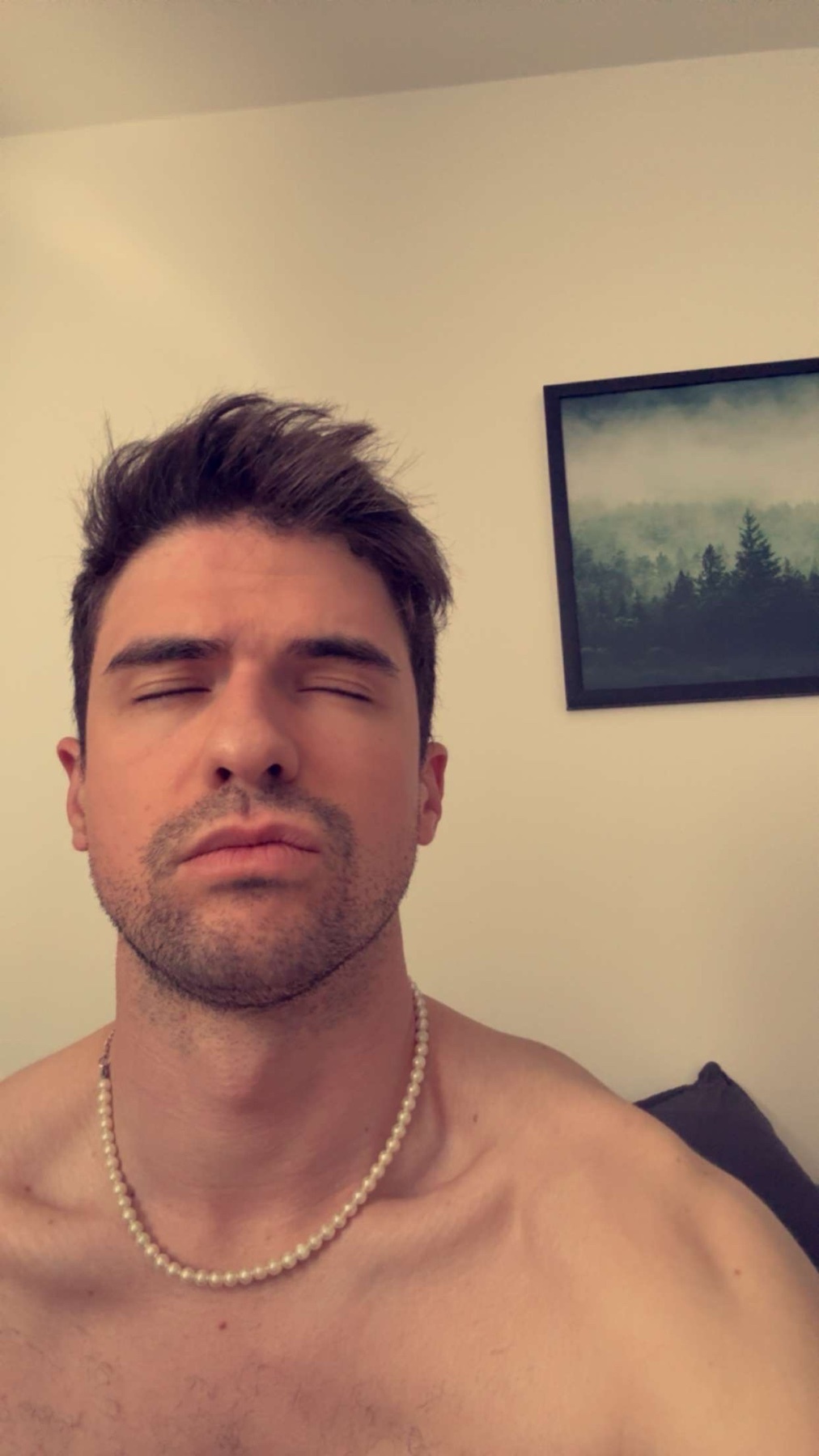 A man with dark hair and stubble is shirtless, wearing a pearl necklace. He has his eyes closed.