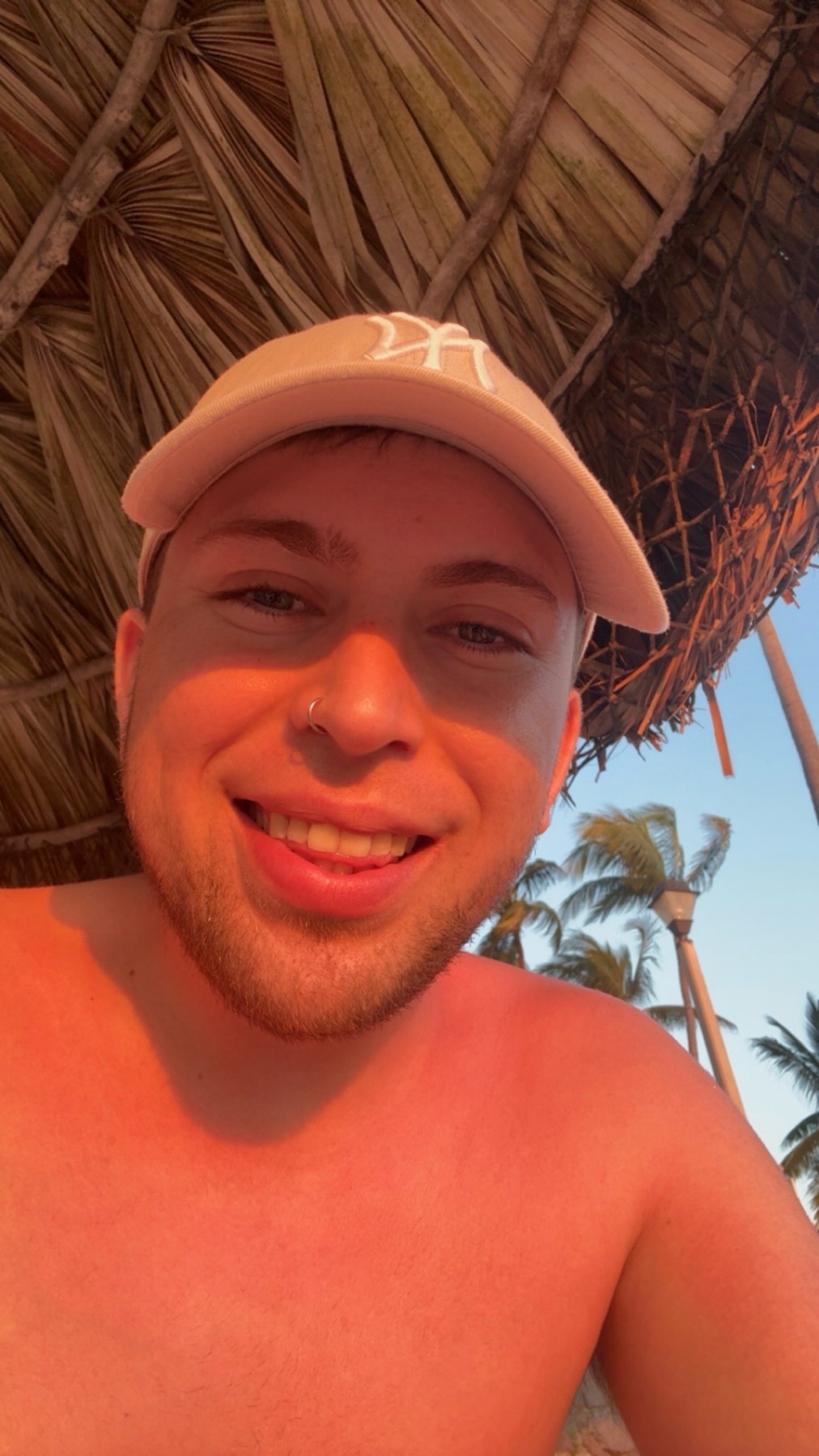 A man wearing a baseball cap and a nose ring smiles at the camera, sticking his tongue out slightly. He is outdoors, with palm trees and a thatched roof visible in the background.
