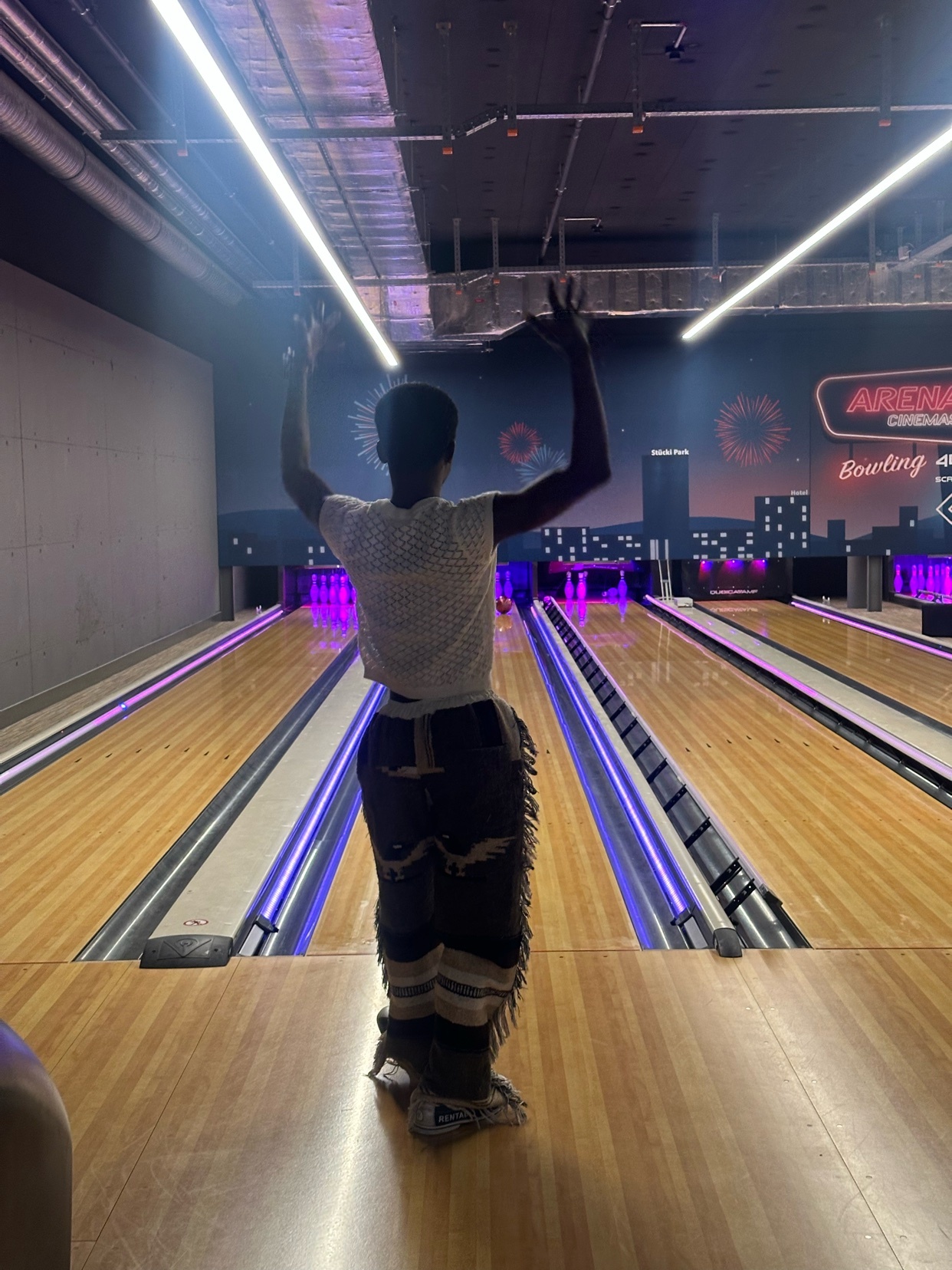 A person stands at the top of a bowling lane, facing away from the camera with their arms raised in celebration. The bowling alley features bright purple lighting and a cityscape mural in the background.
