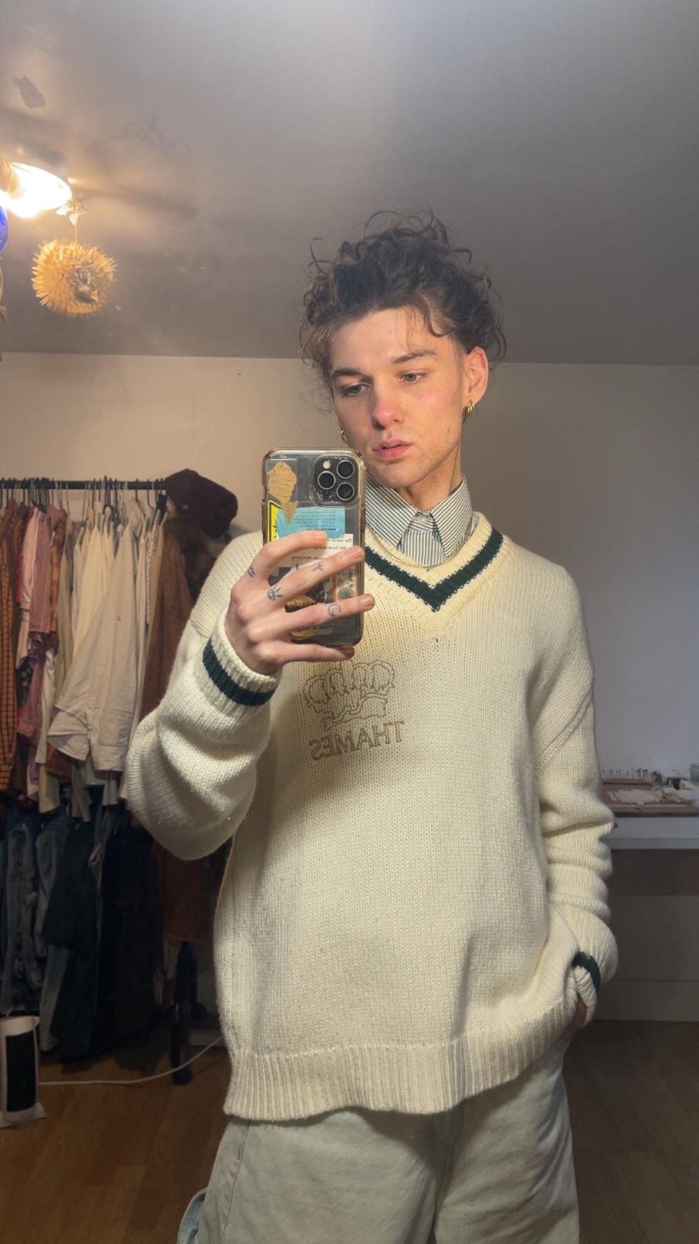 A young man takes a mirror selfie, showcasing his outfit. He's wearing a sweater over a collared shirt, with a modern and stylish aesthetic.