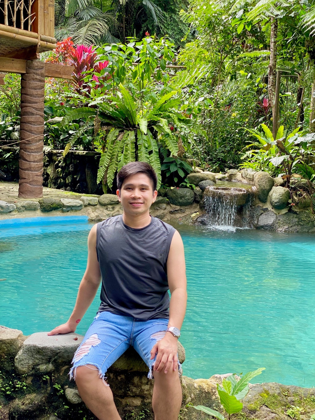 A man poses by a pool in a tropical setting. He is shirtless and wearing shorts.