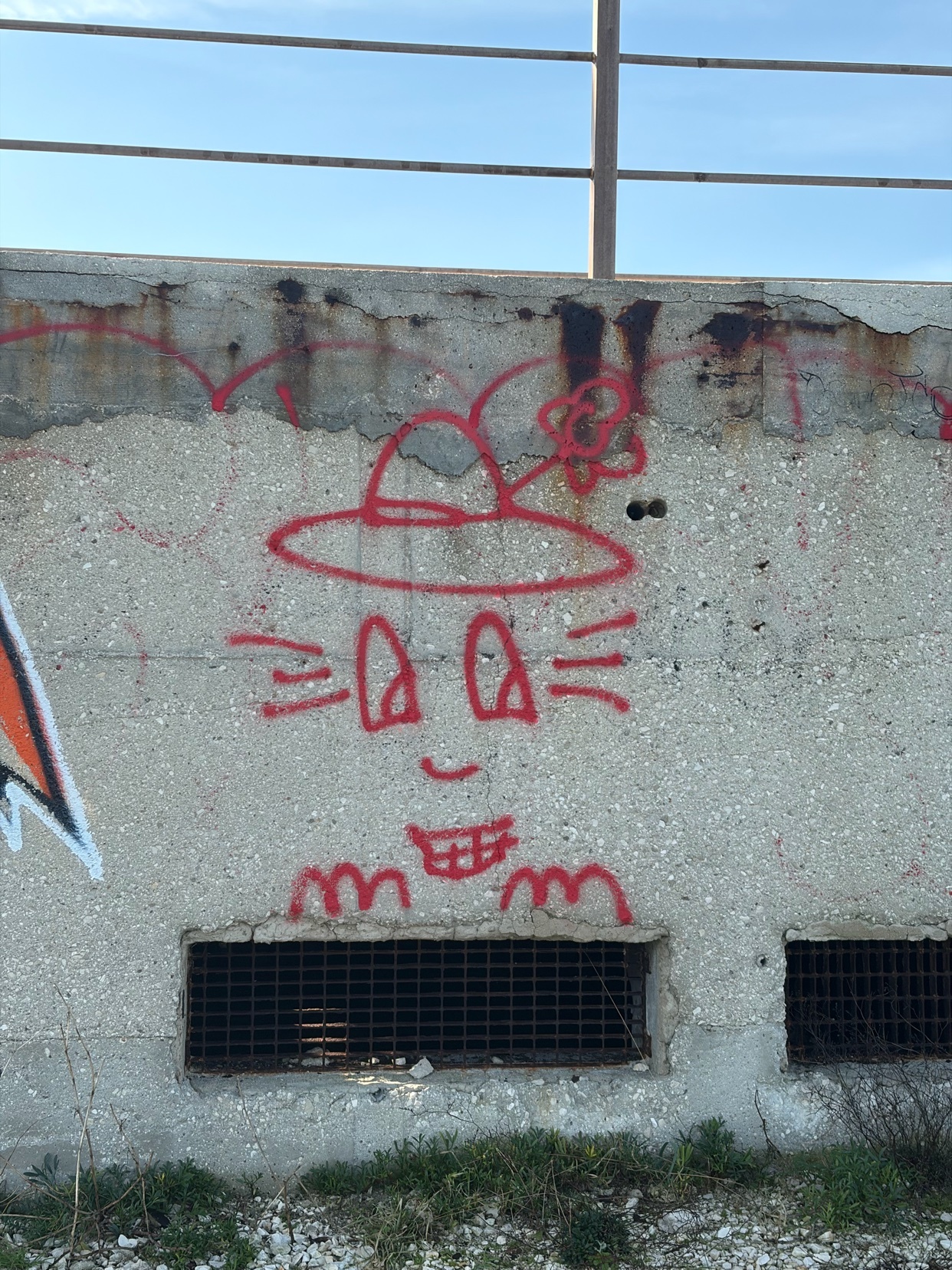 A red graffiti drawing of a face with a hat on a concrete wall. The artwork has a smiling face with big eyes.