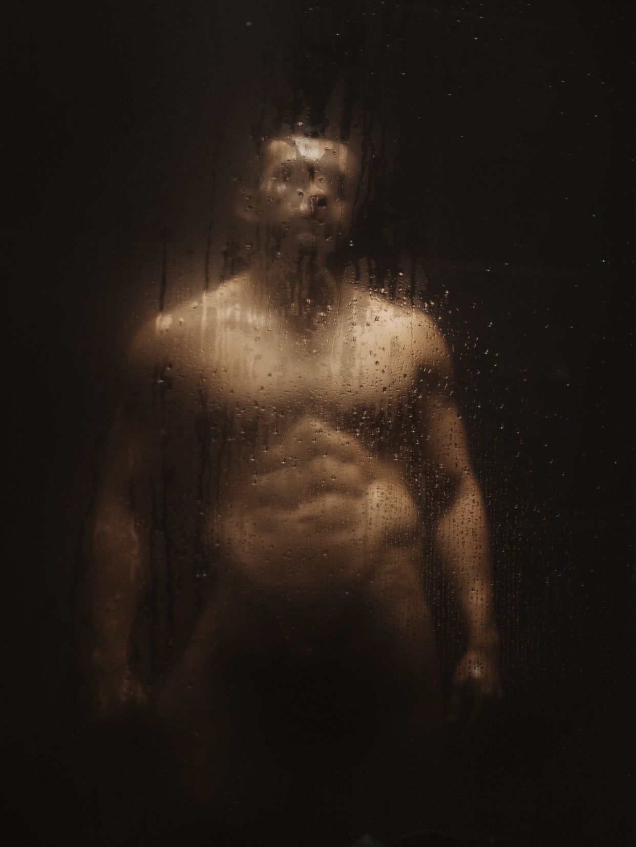A man stands behind a wet, transparent surface, his physique visible through the water droplets. The image has a moody, artistic aesthetic.