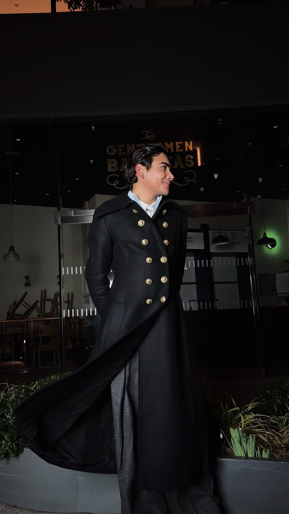 A man poses in a stylish black coat with gold buttons. The photo is taken at night, with a restaurant in the background.
