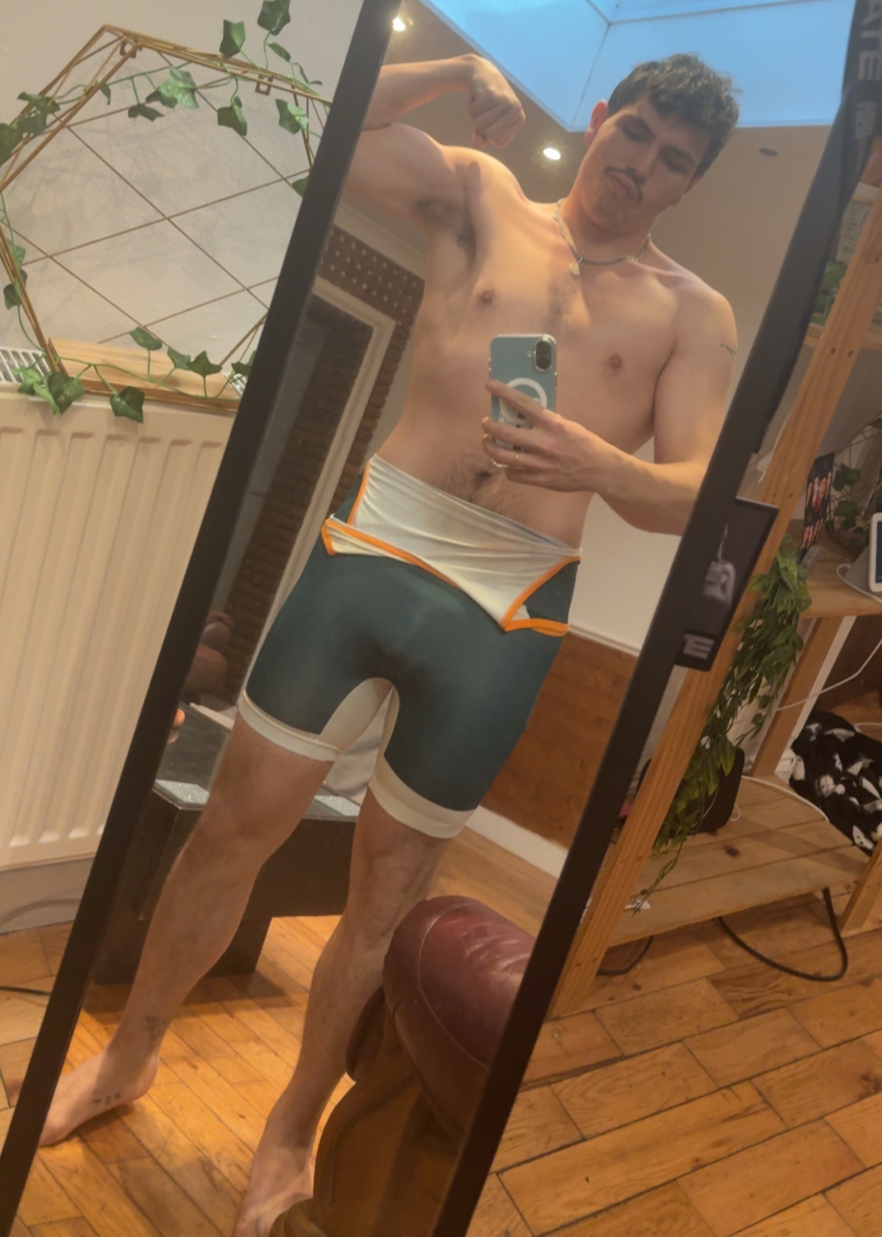 A man takes a mirror selfie, flexing his bicep and showing off his physique in athletic wear. He has stubble and a mustache, and the caption mentions sports gains.