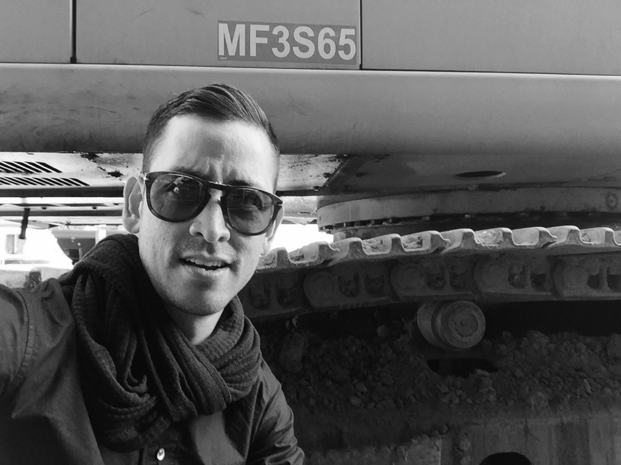 A black and white selfie of a man wearing sunglasses and a scarf. He is standing in front of construction equipment.
