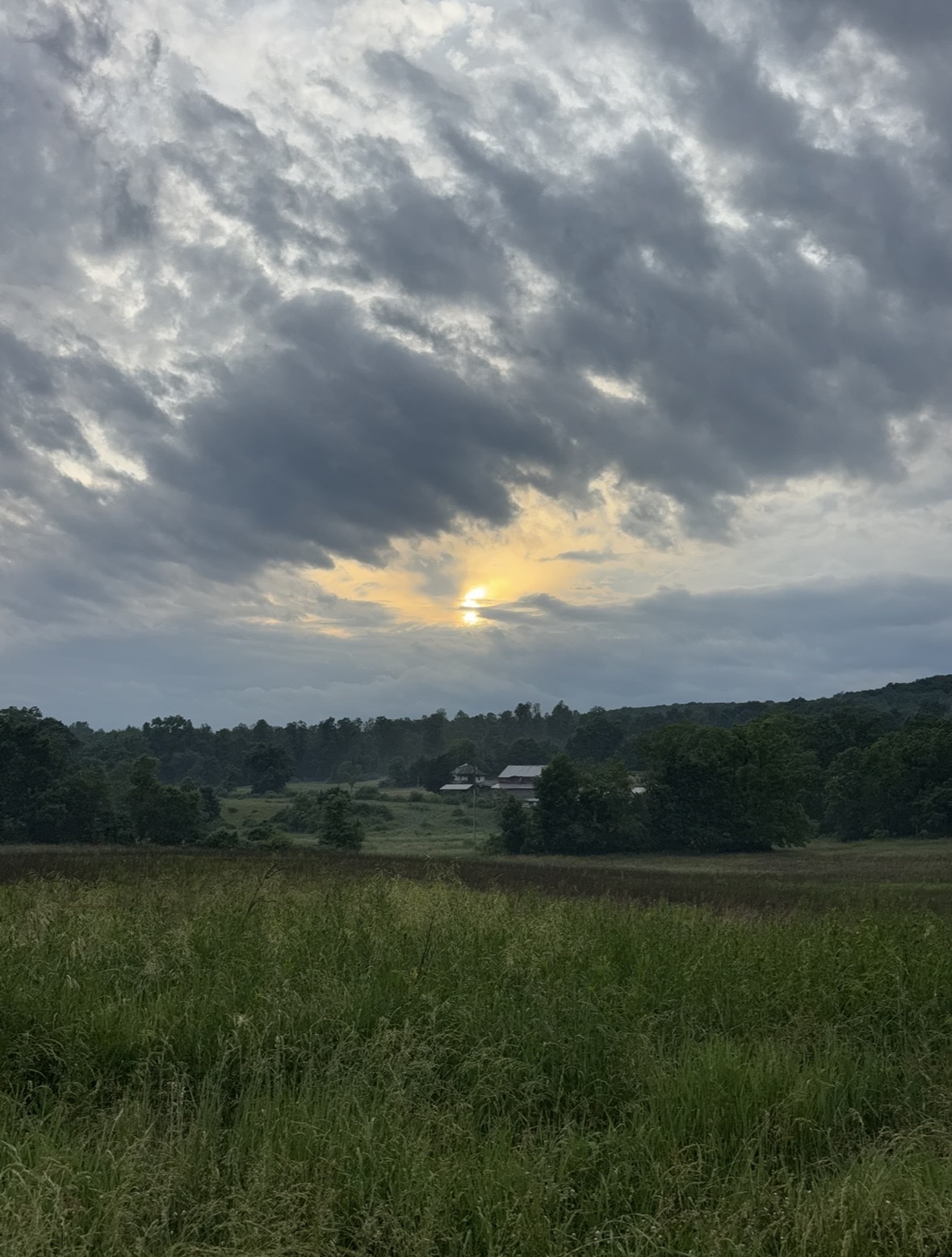 The sun peeks through dramatic, cloudy skies over a rural landscape. A field of tall grass stretches towards a distant farm nestled among trees.