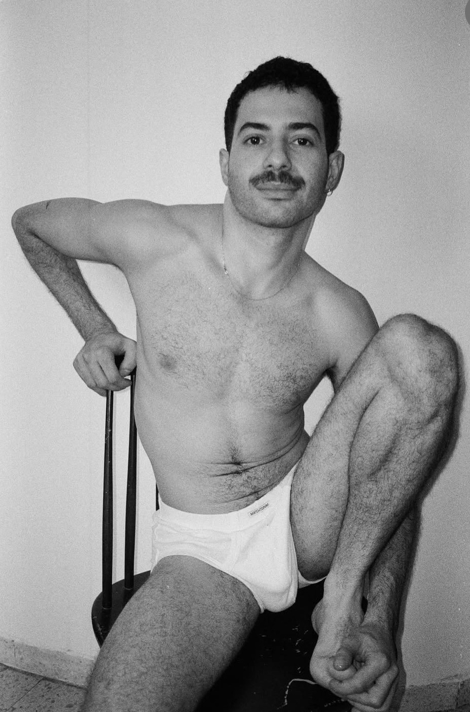 A black and white photo of a man in his underwear, posing on a chair. The man is shirtless and has a mustache.
