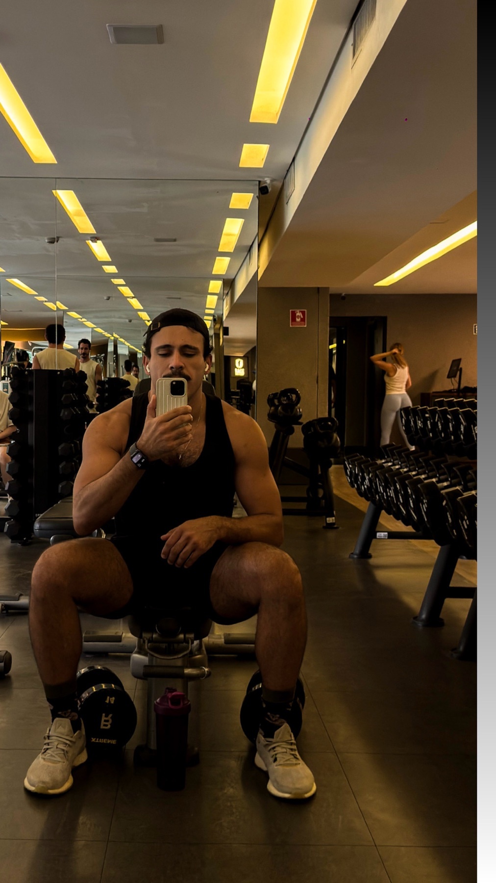 A man in a black tank top and shorts takes a selfie in a gym. He is sitting on a bench with dumbbells and gym equipment in the background.