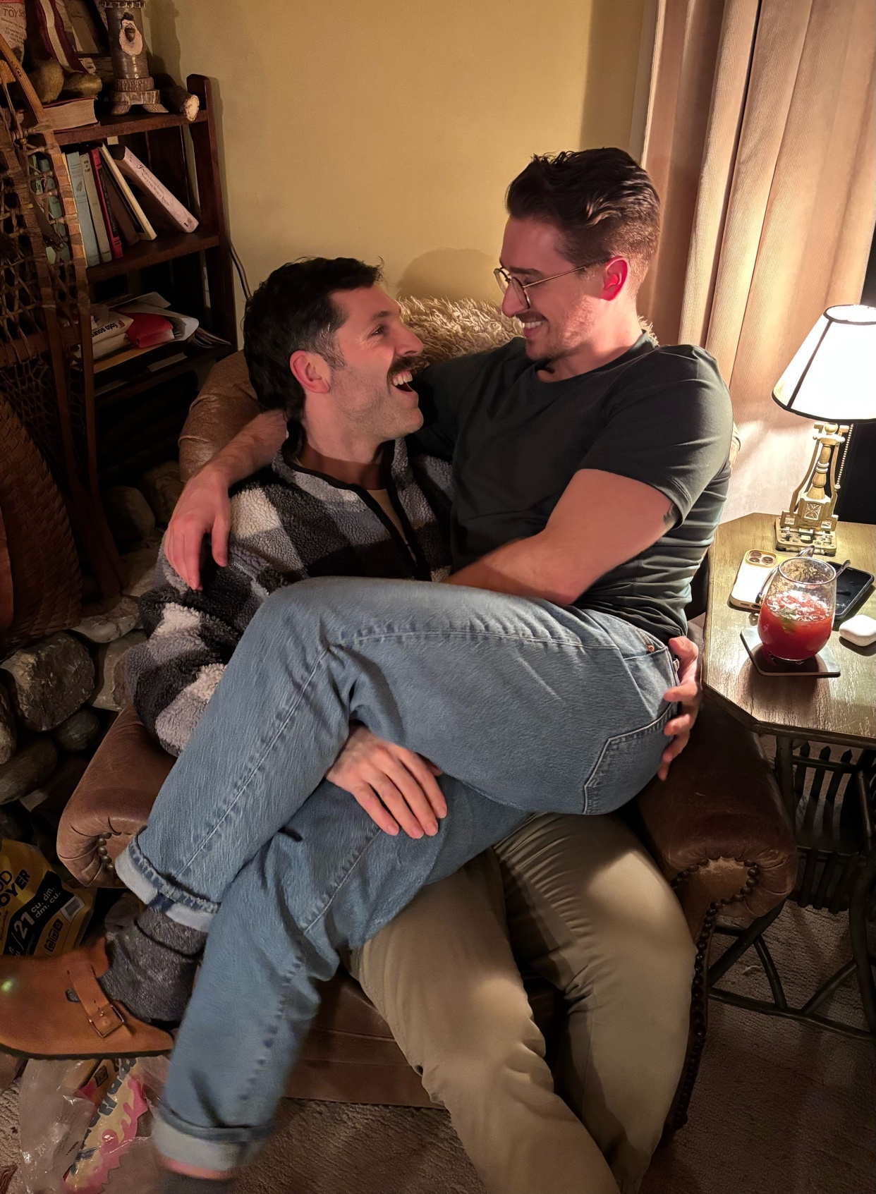 Two men are embracing and smiling at each other. They are sitting in a chair and appear to be in a loving relationship.