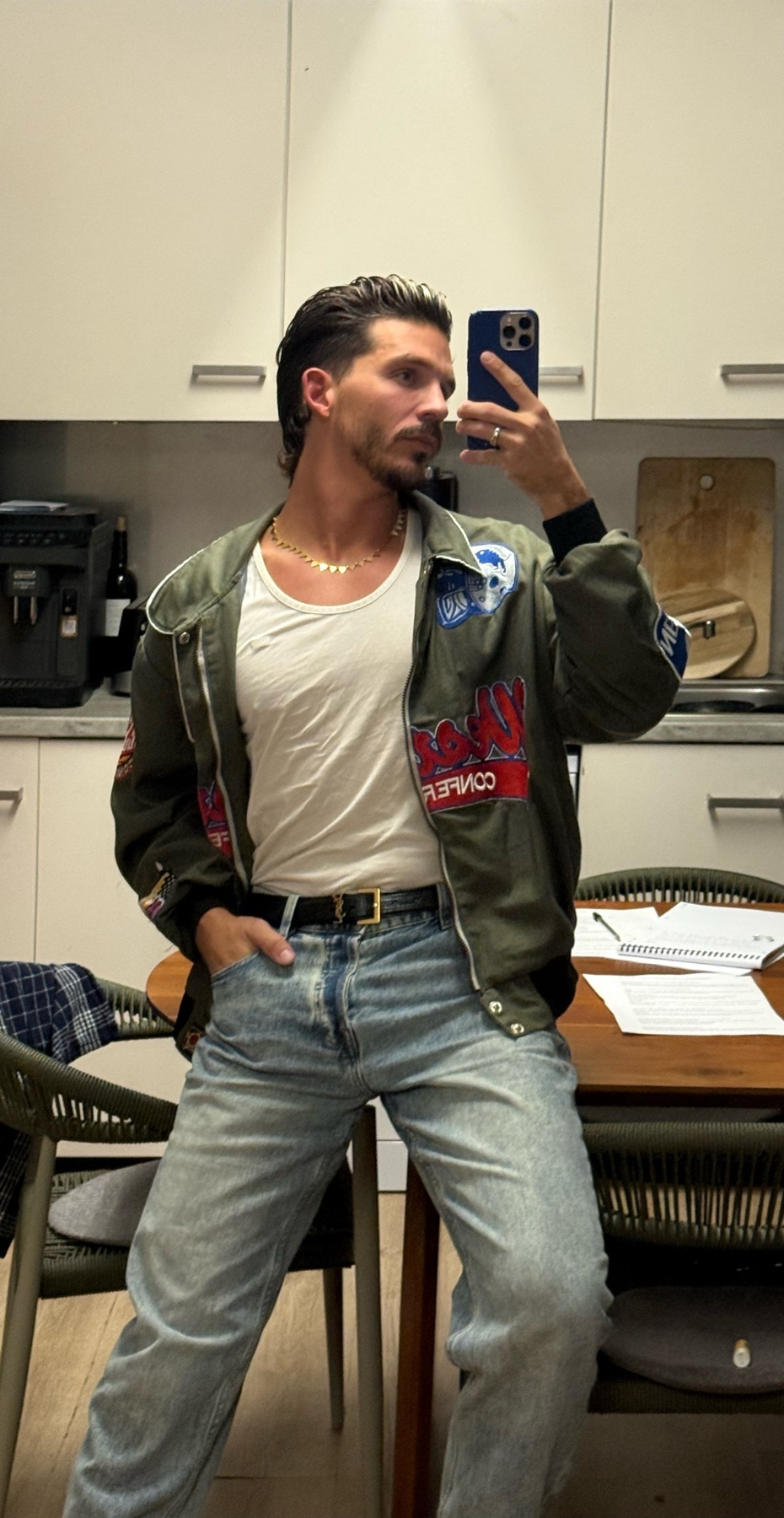 A man takes a mirror selfie in a stylish outfit. He wears a bomber jacket, jeans, and a white t-shirt.