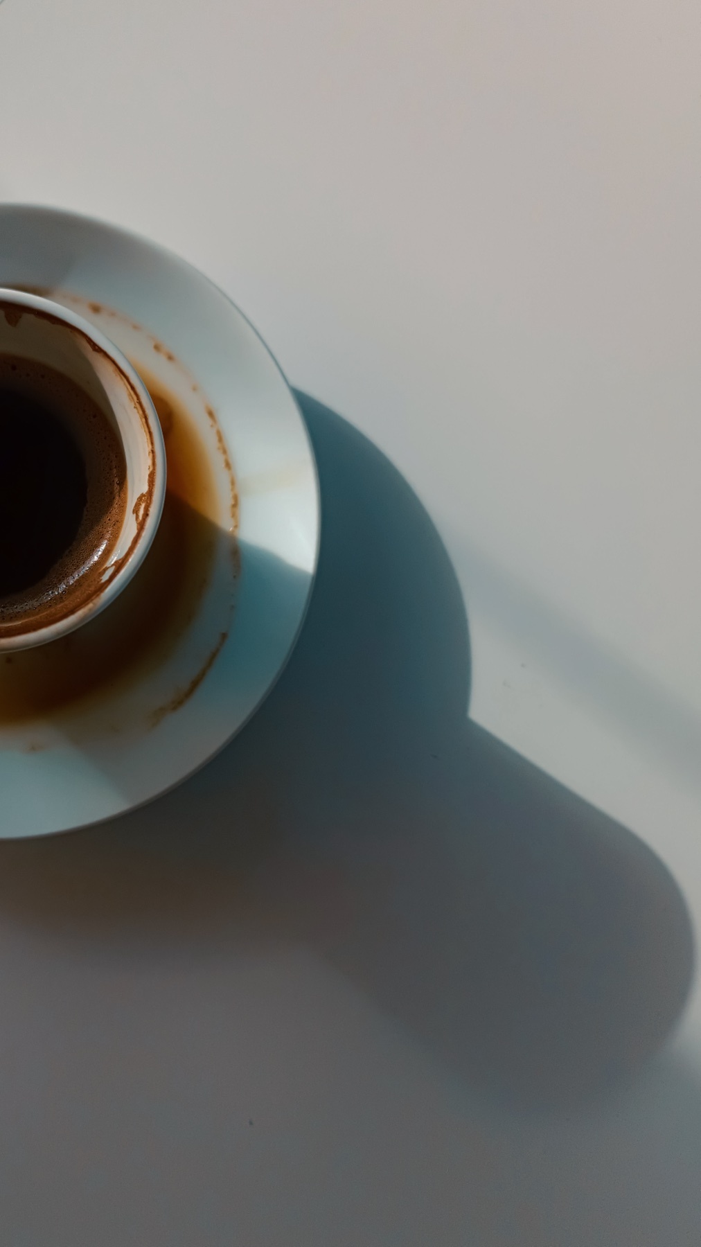 An overhead shot of a coffee cup and saucer, casting a long shadow. The image has a minimalist aesthetic.