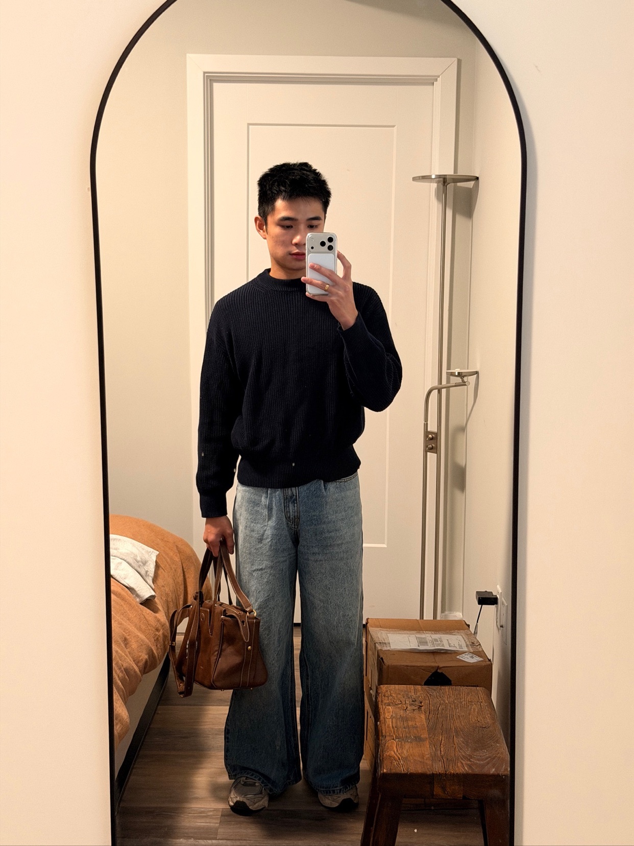 A man takes a mirror selfie, showcasing his outfit. He holds a bag and stands in front of a mirror.
