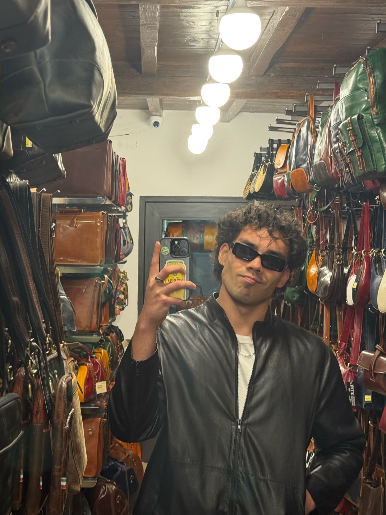 A man with curly hair and sunglasses poses in a shop filled with Italian leather bags.