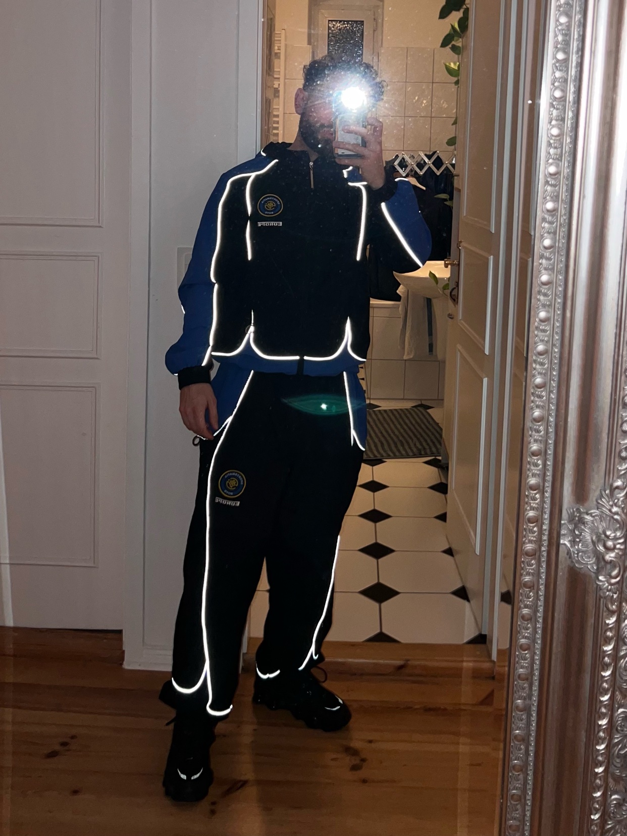 A man poses in a mirror, showcasing a stylish black and blue tracksuit with reflective accents. The outfit is a modern take on streetwear.