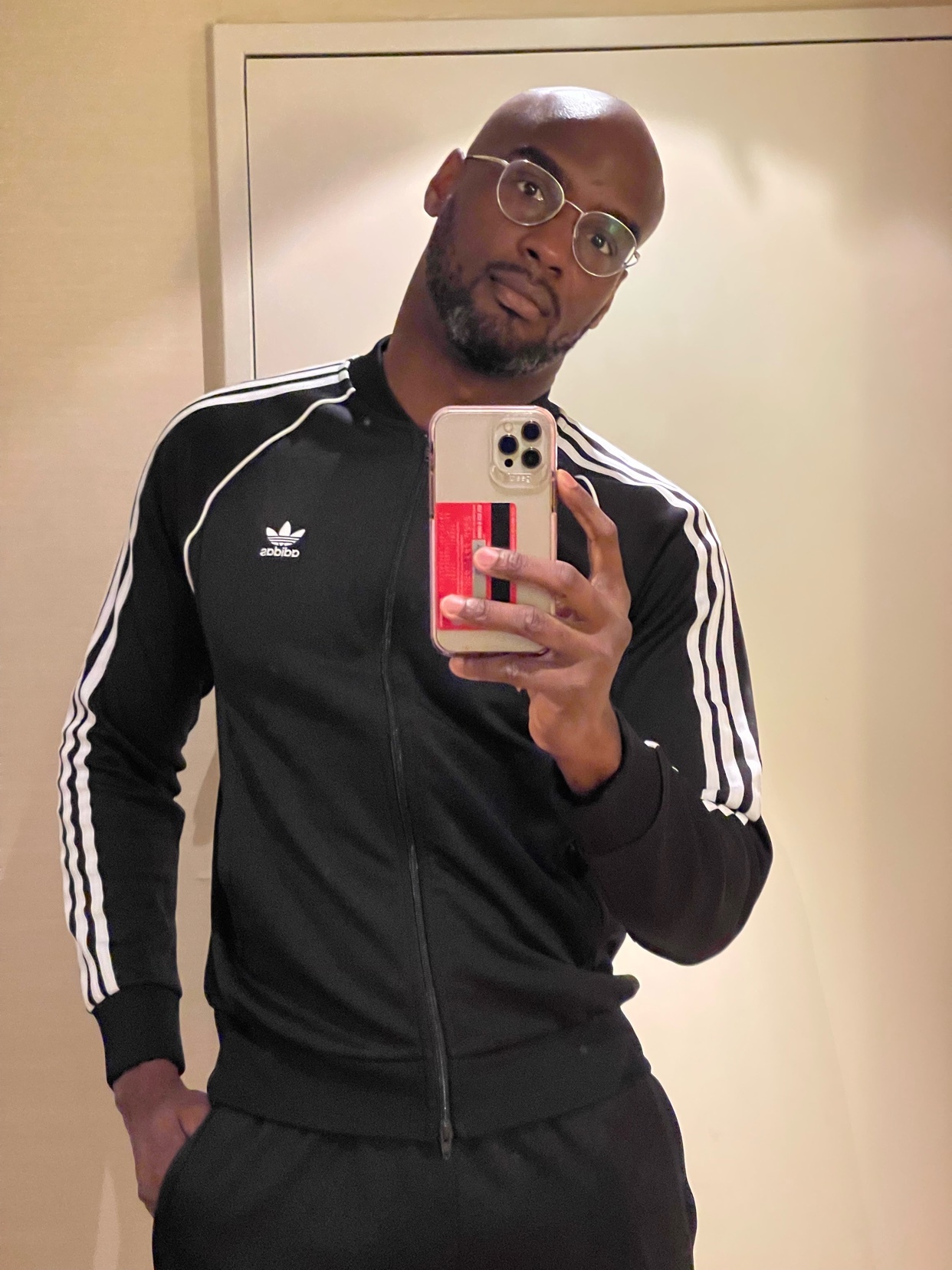 A man takes a mirror selfie, wearing a black Adidas tracksuit and glasses. The photo is taken indoors.