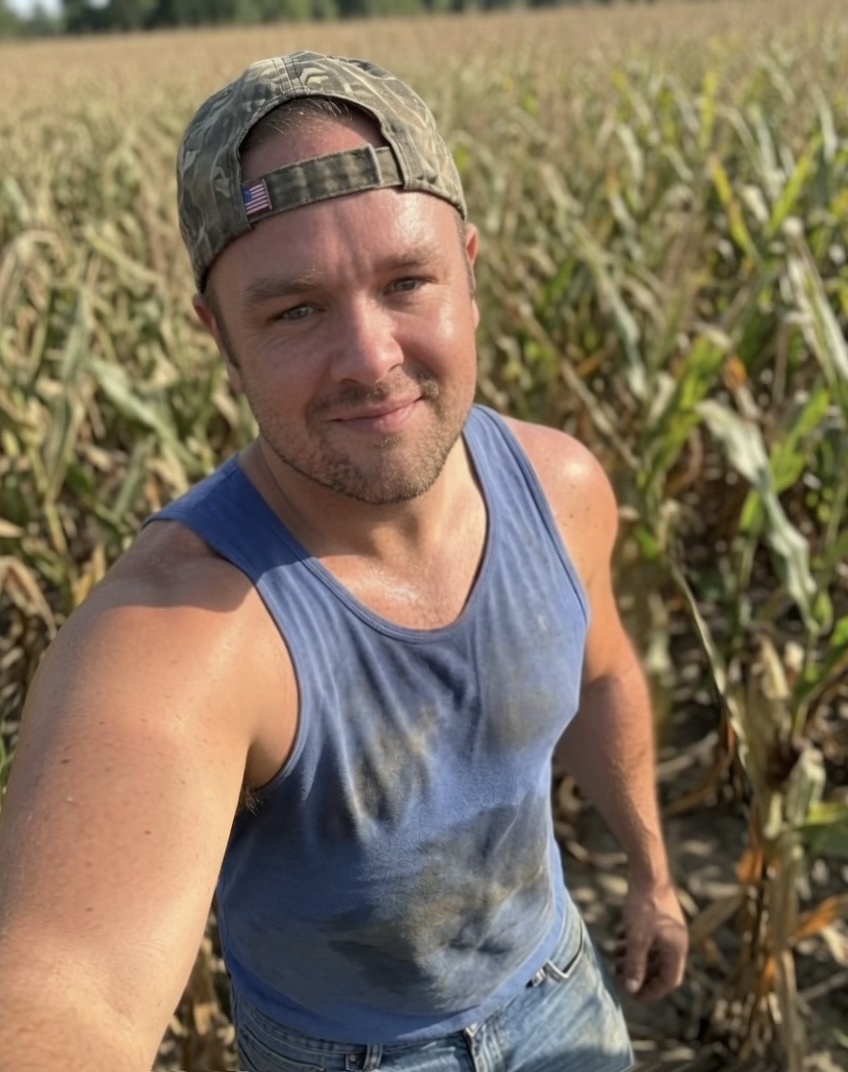 A man in a blue tank top and a camouflage baseball cap takes a selfie in a corn field. He appears to be sweaty and is looking at the camera with a slight smile.