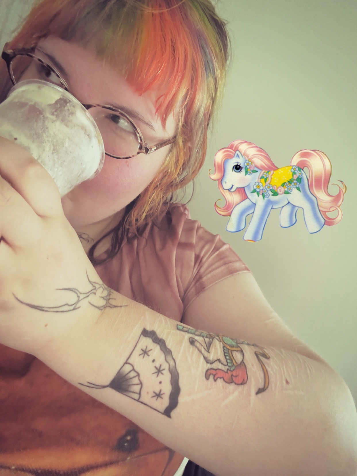 A woman with rainbow-colored hair and glasses drinks from a cup. A vintage My Little Pony graphic is superimposed on the image.