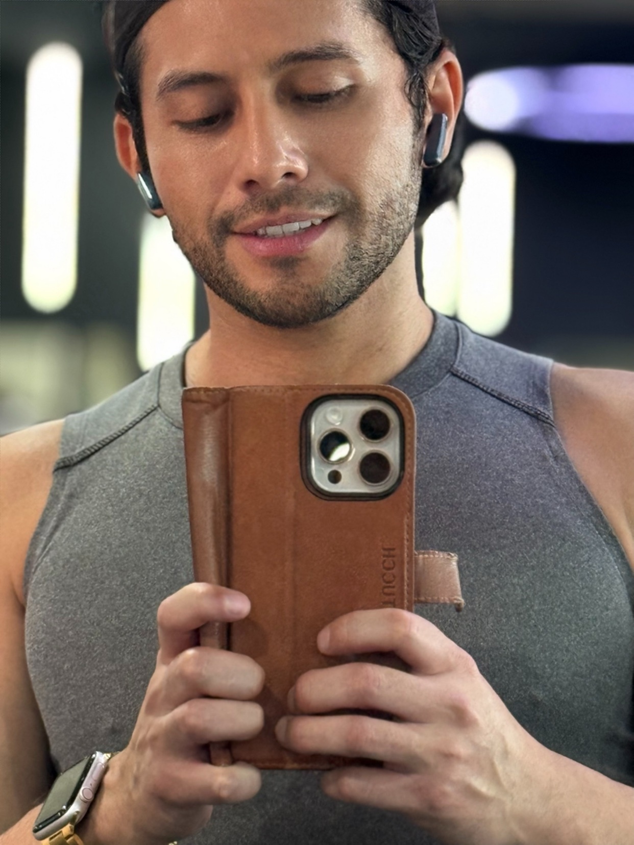 A man takes a selfie in a gym, holding his phone in a brown leather case. He is wearing wireless earbuds and a grey tank top.