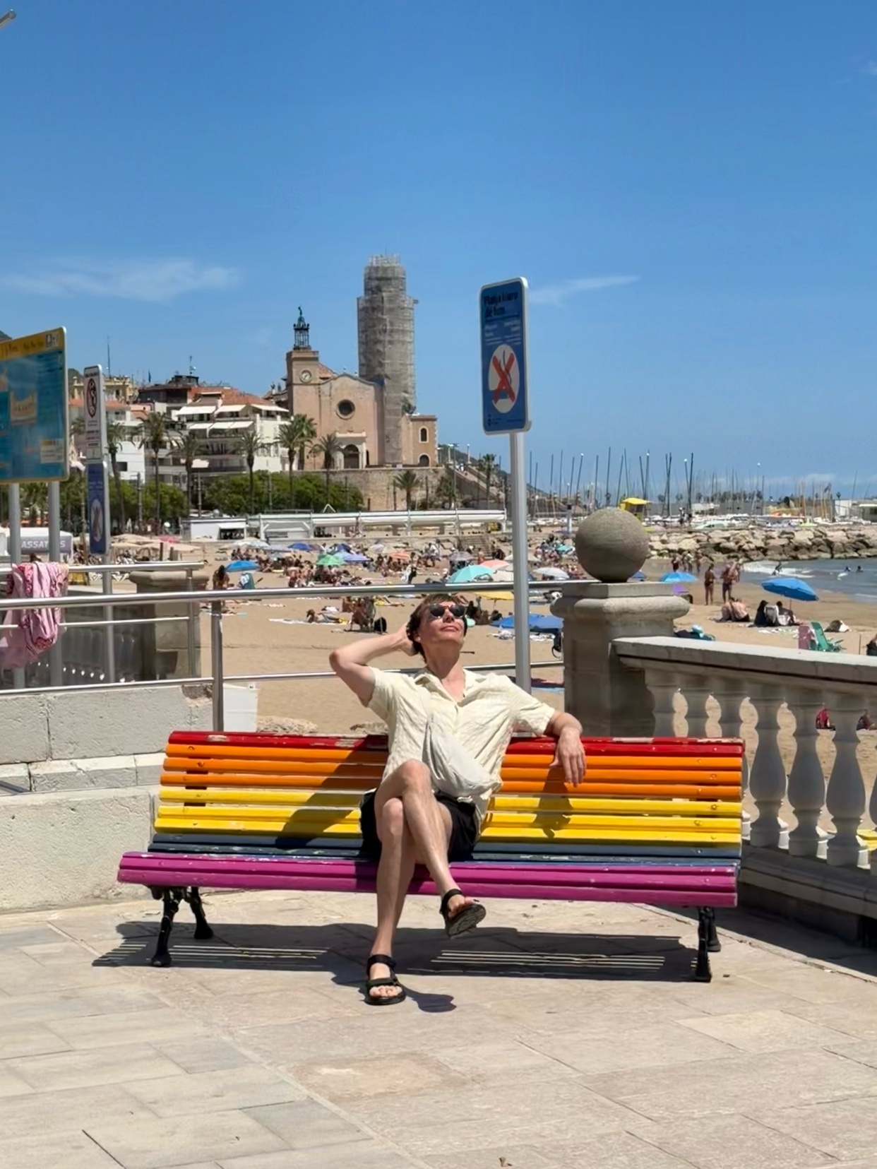 A man sits on a rainbow-colored bench overlooking a crowded beach on a sunny day. The man is wearing sunglasses and a short-sleeved shirt, with his head tilted back and one hand behind his head.
