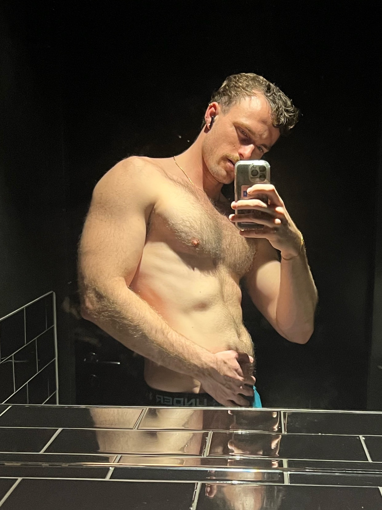 A man takes a mirror selfie, showcasing his physique. He is shirtless, flexing his bicep.