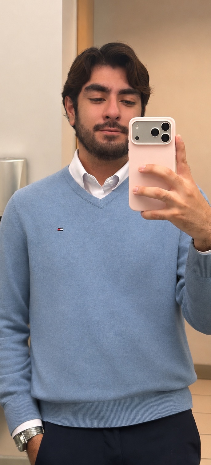 A man takes a selfie in a light blue v-neck sweater over a white button-down shirt. He is wearing a watch on his left wrist and has a beard and dark hair.
